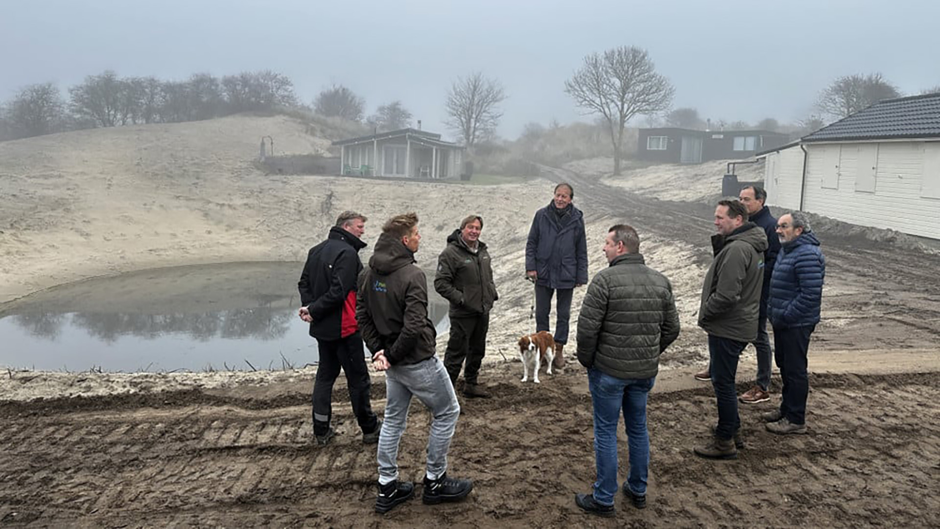Een groep mensen staat buiten in een zandachtig duinlandschap met een vijver en enkele gebouwen op de achtergrond. Een hond is ook aanwezig.