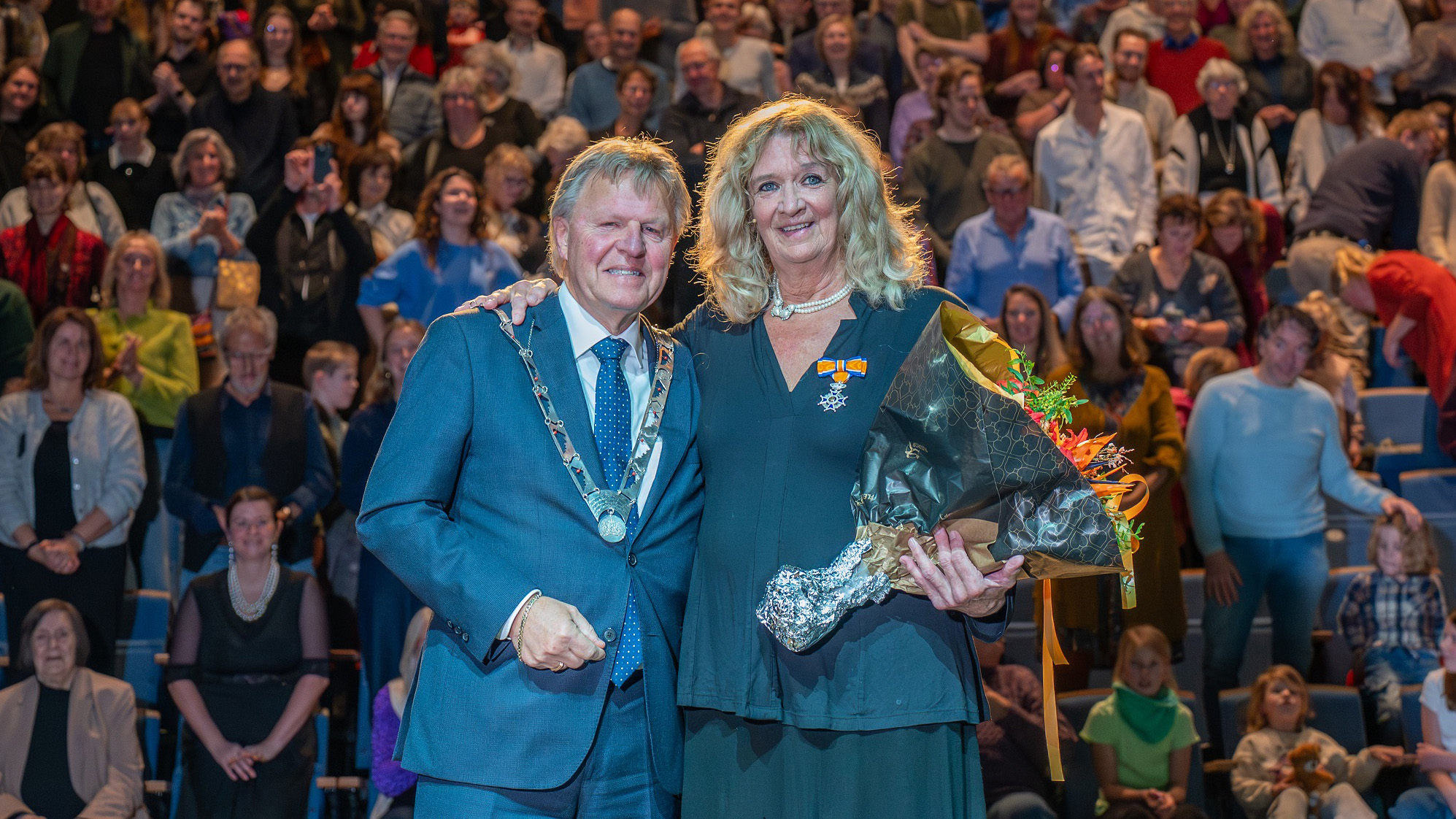 Twee personen poseren voor een volle zaal; een man in een blauw pak met ambtsketen en een vrouw met een bloemenboeket en onderscheidingslint op haar kleding.