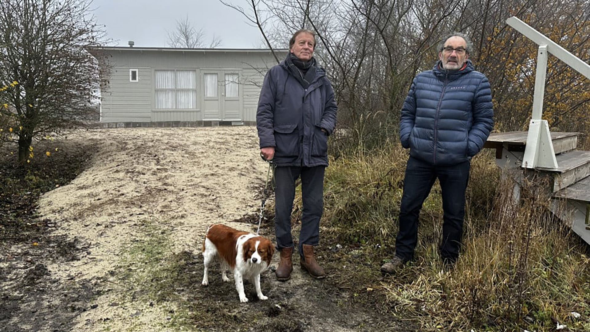 Twee mannen staan buiten naast een hond aan de lijn voor een grijs vakantiehuis, omgeven door kale bomen en struiken.