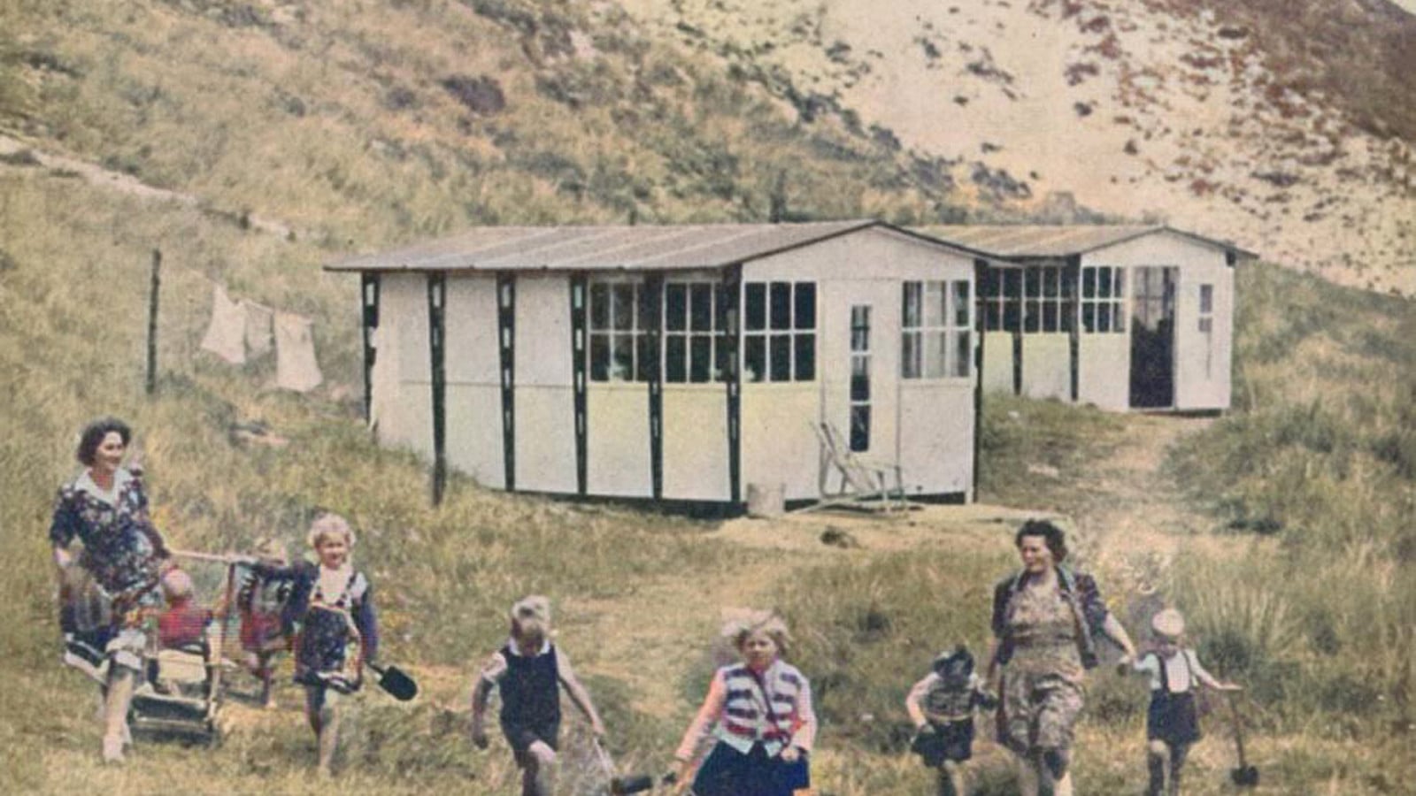 Een groep vrouwen en kinderen loopt in een grasrijk gebied voor een eenvoudige witgeschilderde barak met ramen. Op de achtergrond hangen kleren aan een waslijn naast de barak.