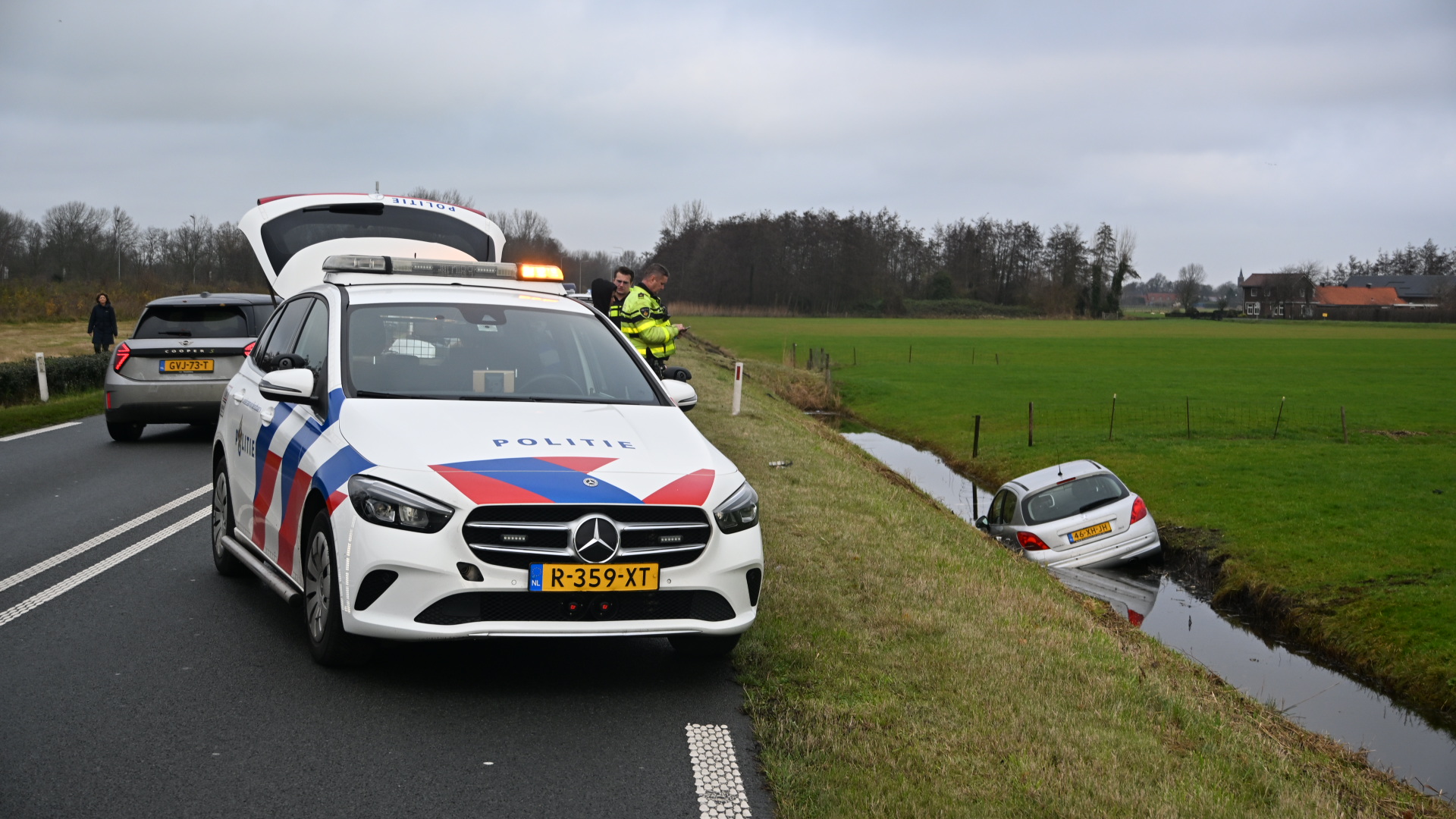 Een politieauto met een geactiveerd zwaailicht staat langs een weg naast een greppel, waarin een zilveren auto gedeeltelijk in het water staat. Twee politieagenten staan naast de politieauto en een persoon loopt naar een andere auto op de weg. Op de achtergrond is een veld en enkele gebouwen te zien.