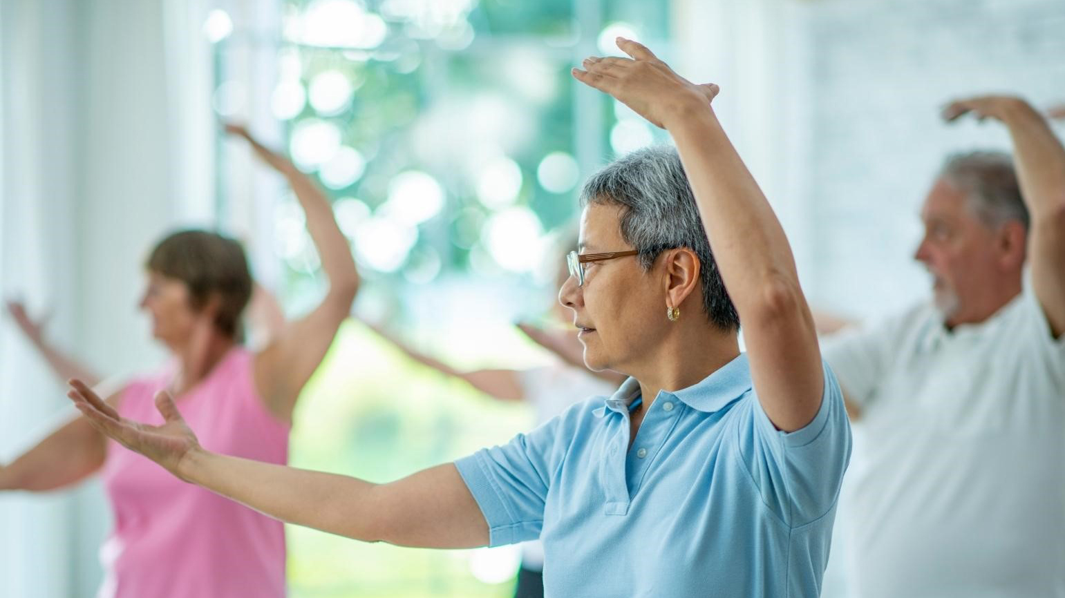 Een groep oudere volwassenen doet Tai Chi-oefeningen in een lichte ruimte, waarbij een vrouw een blauwe blouse draagt en gefocust naar voren kijkt.