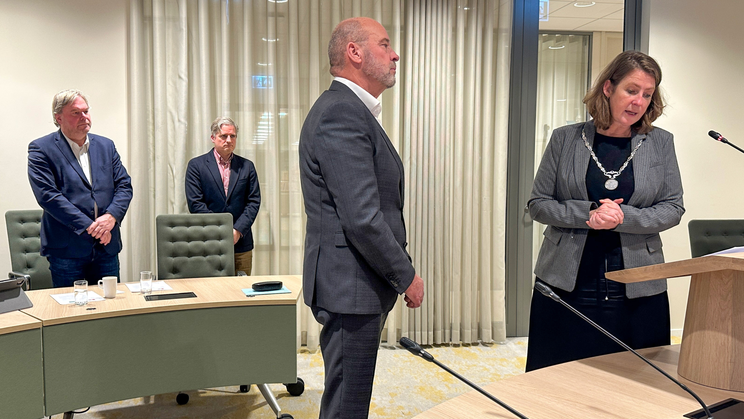 Een man in pak staat tegenover een vrouw in een grijze blazer die achter een lessenaar staat in een vergaderruimte. Ze draagt een keten en spreekt in een microfoon. Twee andere mannen in pakken staan op de achtergrond bij een ronde vergadertafel.