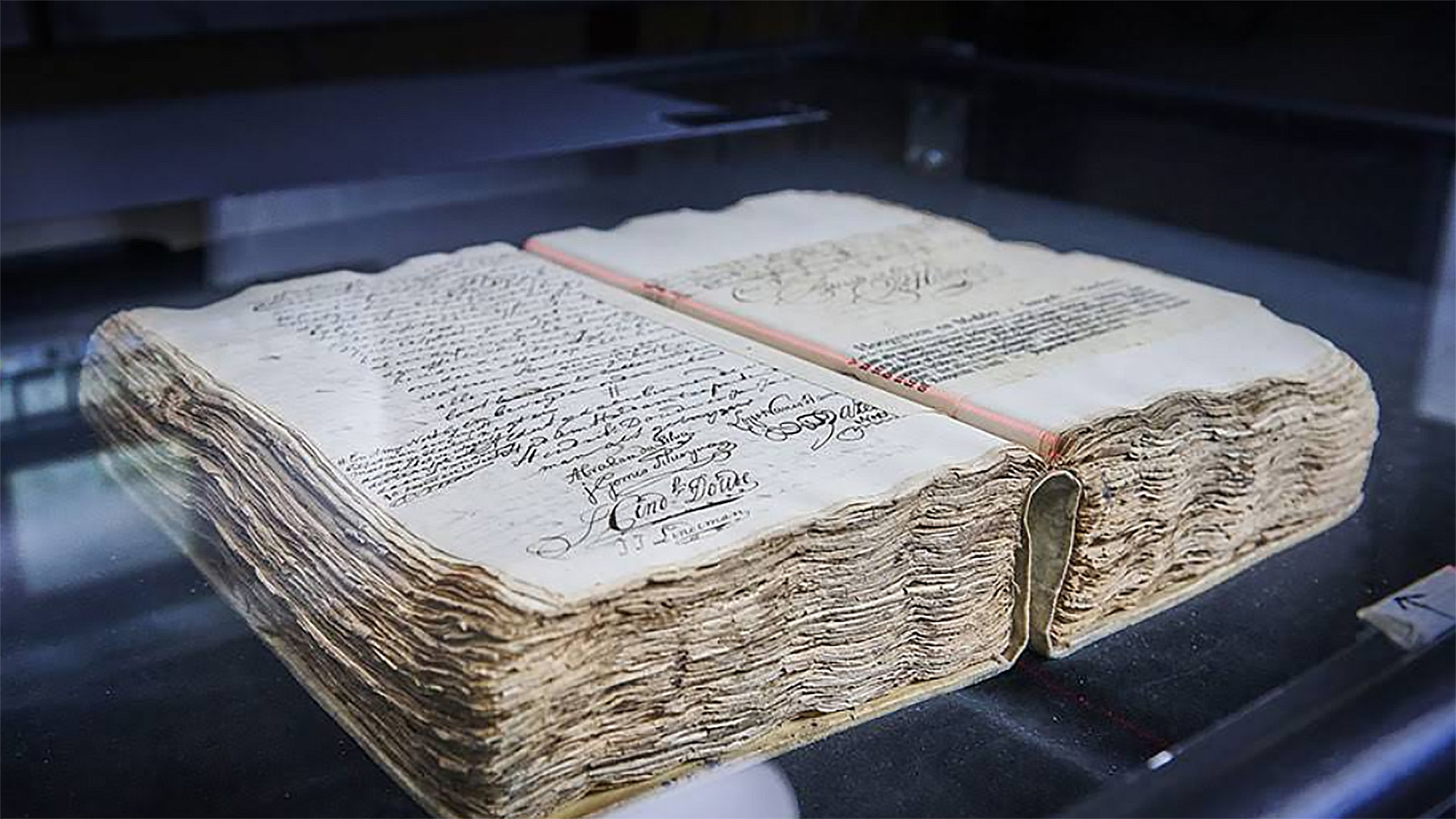 Een oud, dik boek met handgeschreven tekst en versierde handtekeningen op de pagina's, vertoond onder een glazen dekking.