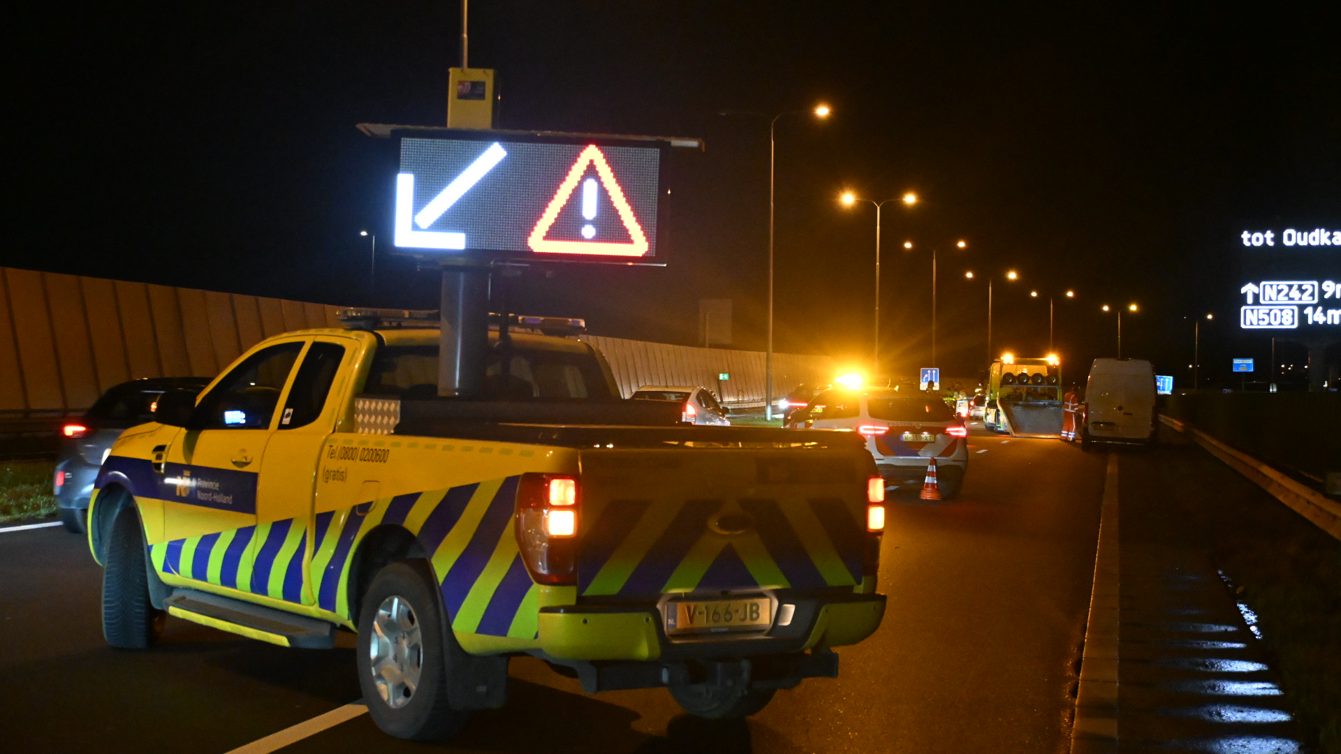 Een gele hulpdienstenwagen met waarschuwingslichten staat stil op de snelweg bij nacht, met verkeer dat eromheen rijdt en verkeersborden zichtbaar op de achtergrond.