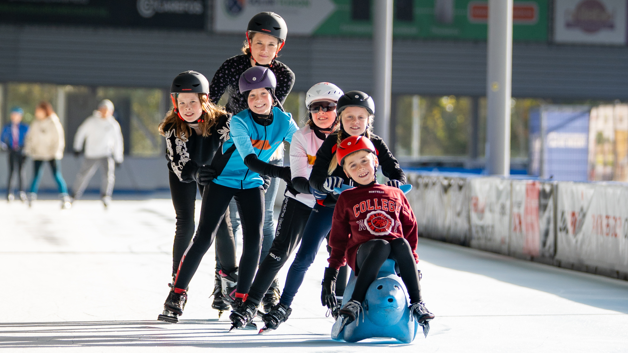 Groep kinderen in winterkleding en helmen schaatst in een rij op een ijsbaan, met een kind dat voorop zit op een blauwe slee. Ze lachen en hebben plezier.