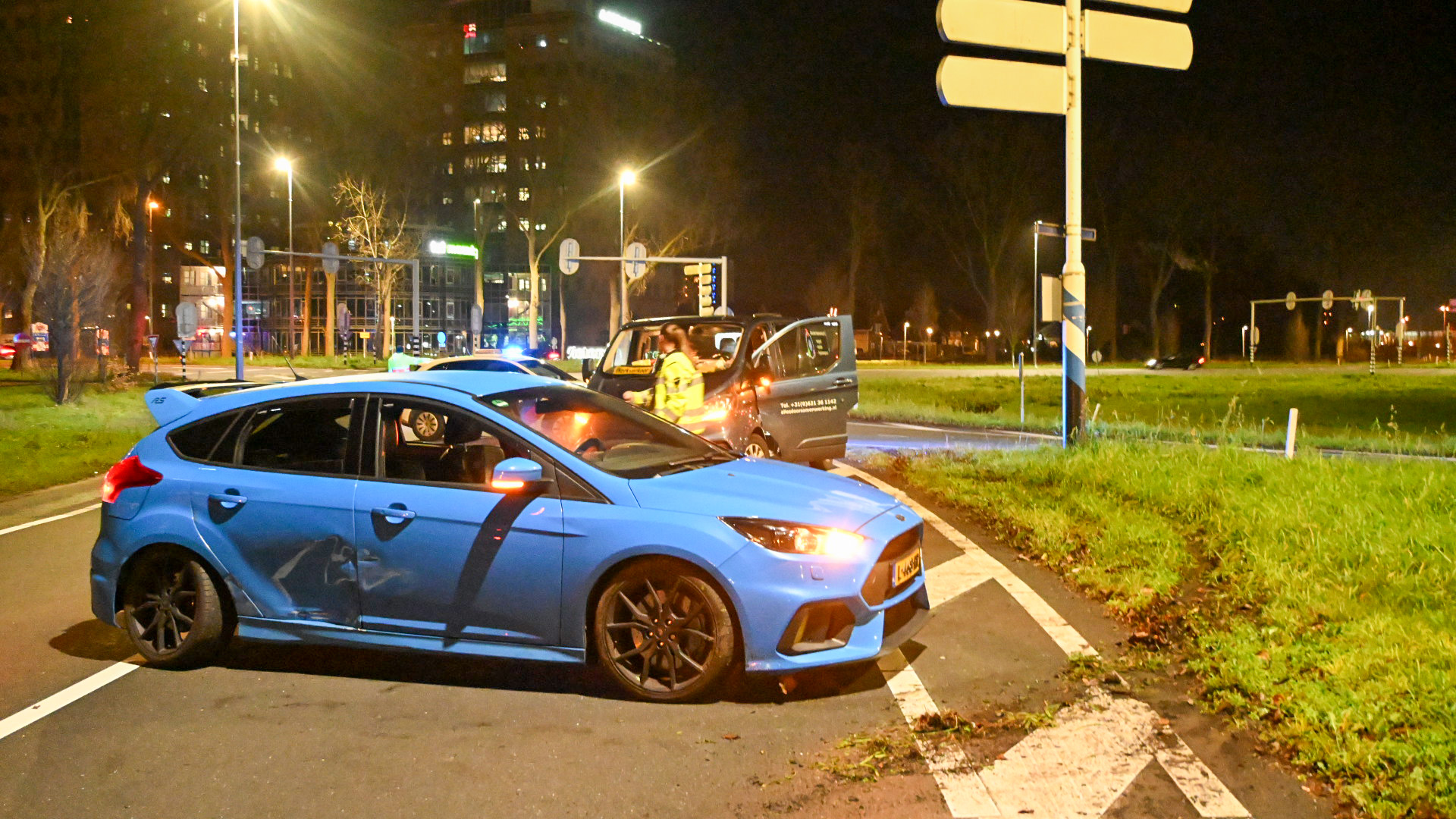 Een blauwe auto met schade aan de zijkant en een zwarte taxi zijn betrokken bij een botsing op een rotonde, verlicht door straatlantaarns, met op de achtergrond een gebouw en een stuk gras.