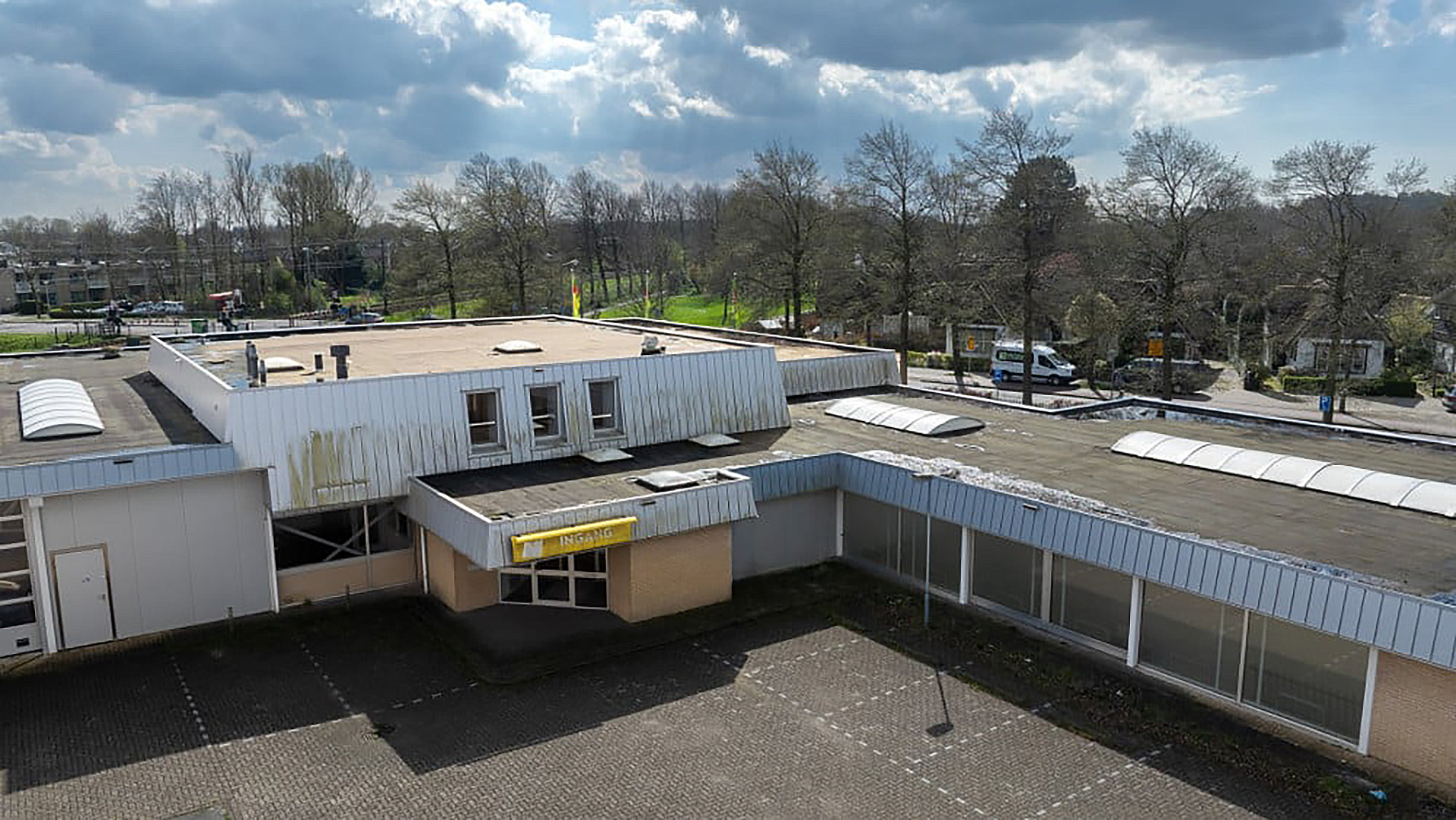 Luchtfoto van een verouderd gebouw met een plat dak en enkele ramen, omringd door een lege parkeerplaats en kale bomen onder een bewolkte lucht.