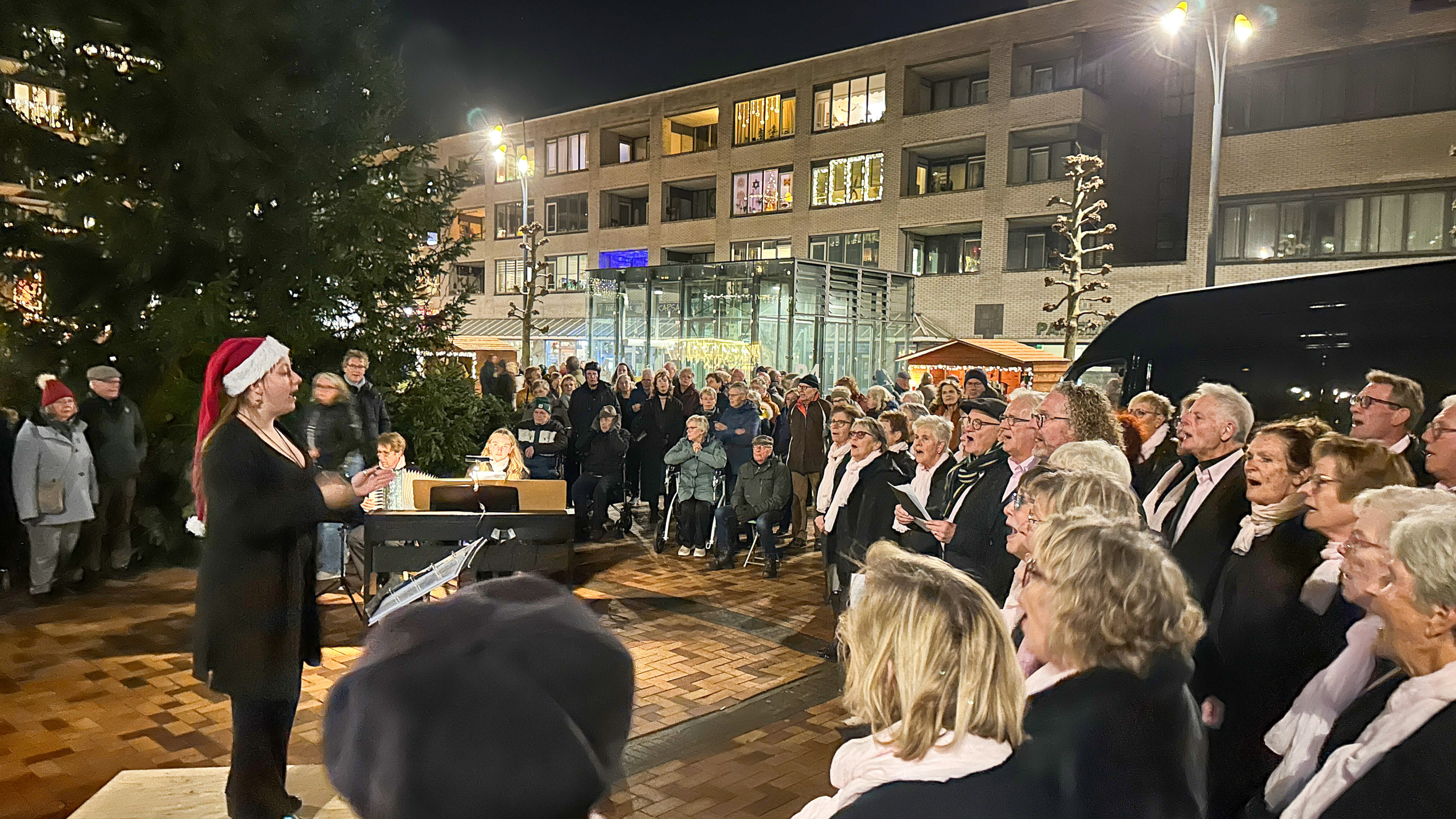 Een kerstkoor zingt in een openluchtplek 's avonds, geleid door een dirigent met een kerstmuts, omringd door een menigte van toeschouwers.