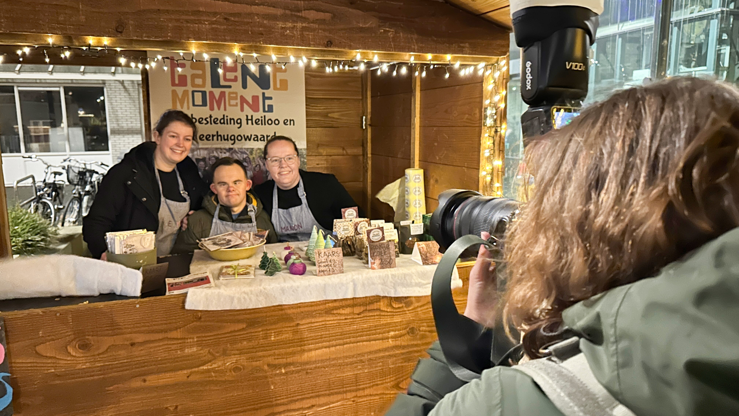 Drie mensen poseren voor een kerstkraam met versierde kaarsen en koekjes, terwijl een fotograaf met een camera een foto maakt.