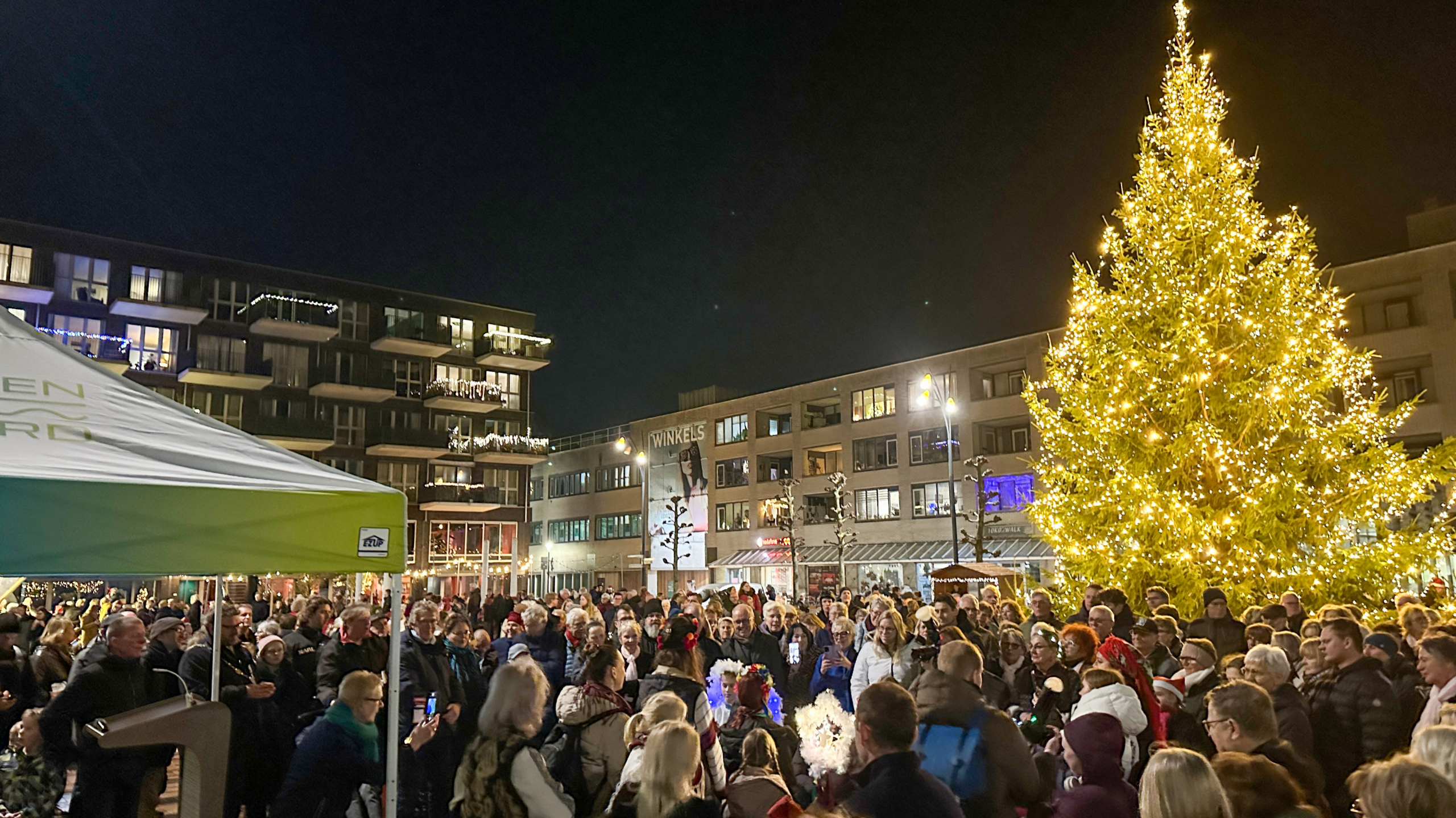 Mensen verzamelen zich 's avonds rond een grote, verlichte kerstboom op een plein, met een tent op de voorgrond en gebouwen met kerstverlichting op de achtergrond.