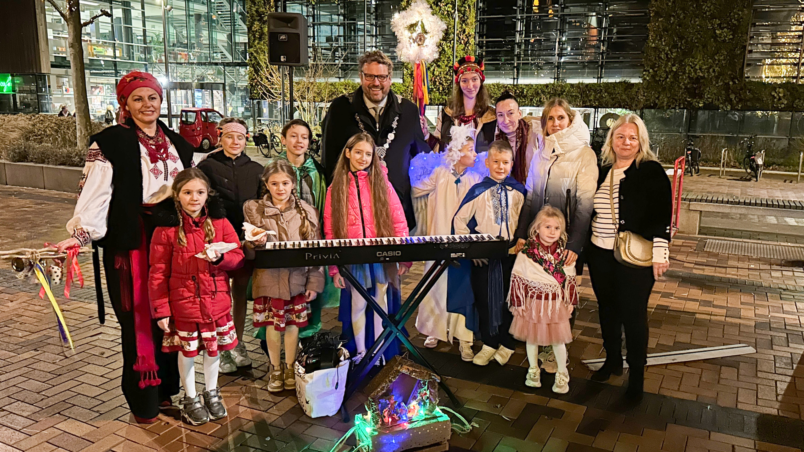 Een groep vrouwen en kinderen, sommigen gekleed in traditionele kleding, poseren 's avonds buiten rond een keyboard en een miniatuur kersttafereel, met een gebouw op de achtergrond.