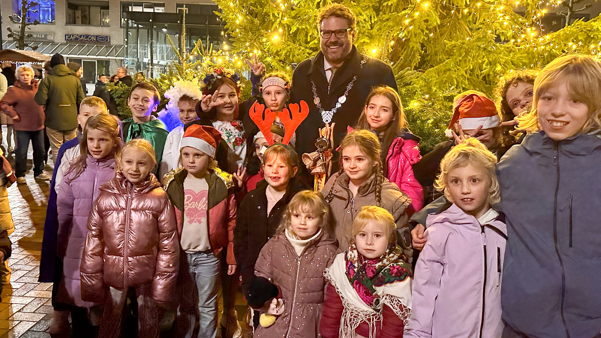 Een groep kinderen poseert naast een kerstboom met feestverlichting aan, vergezeld door een volwassen man met een ketting om zijn nek. Enkele kinderen dragen kerstmutsen en rendiergeweien.