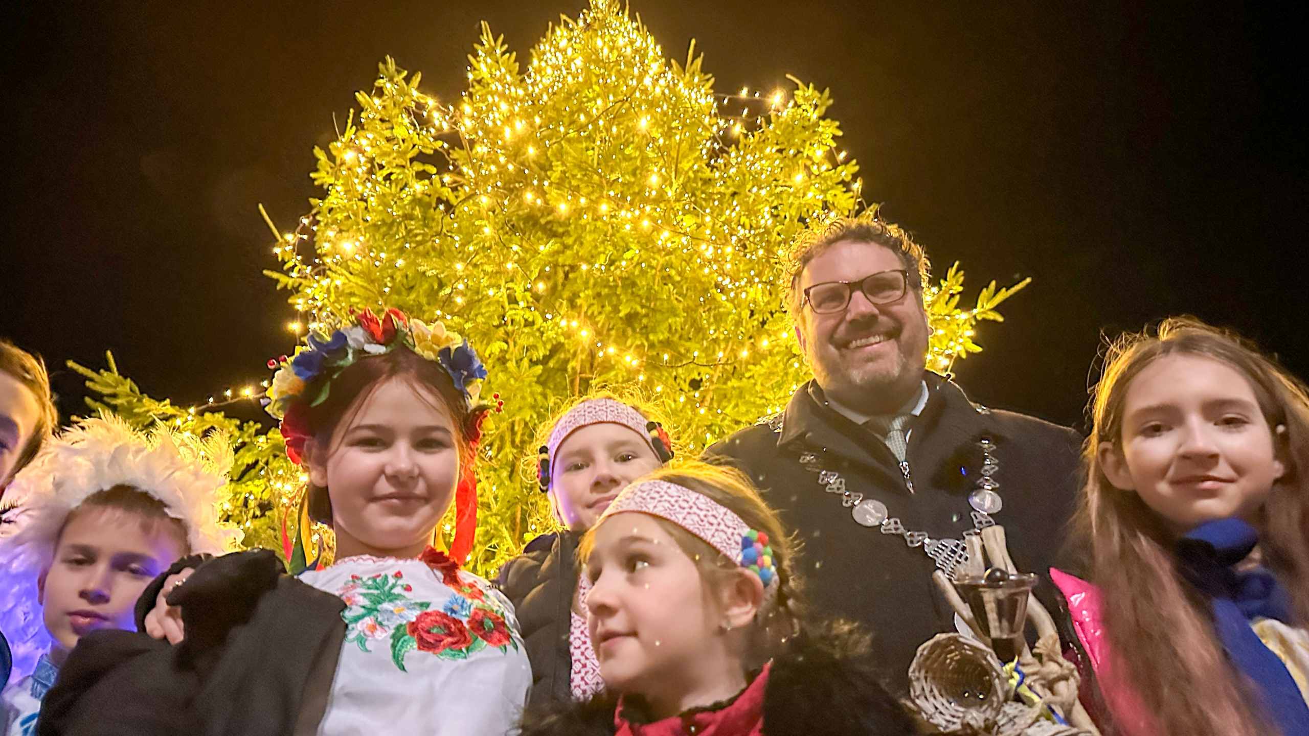 Groep mensen, waaronder kinderen in traditionele kleding, poseert voor een versierde kerstboom met kerstlichtjes in de avond.