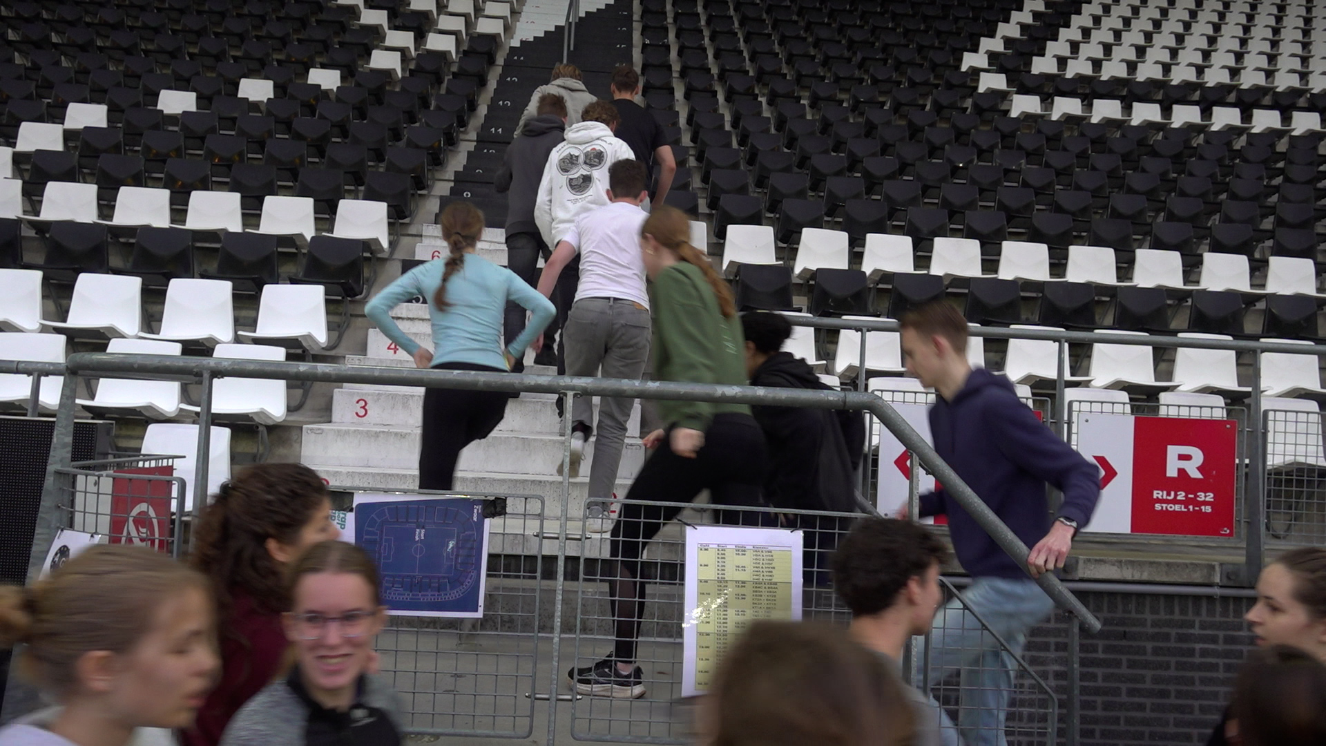 Een groep mensen rent een trap op in een leeg stadion met zwart-witte stoelen, naast een hek met informatieborden.
