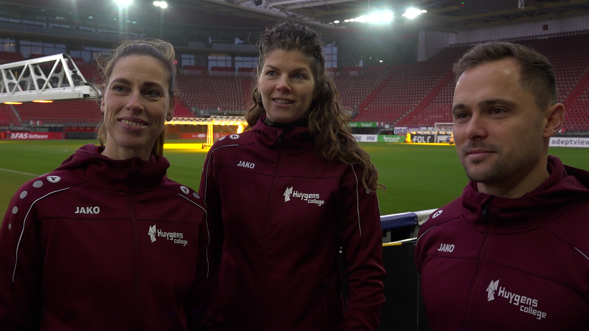 Drie mensen in trainingsjacks met 'Huygens College' logo staan in een stadion met lege tribunes op de achtergrond.