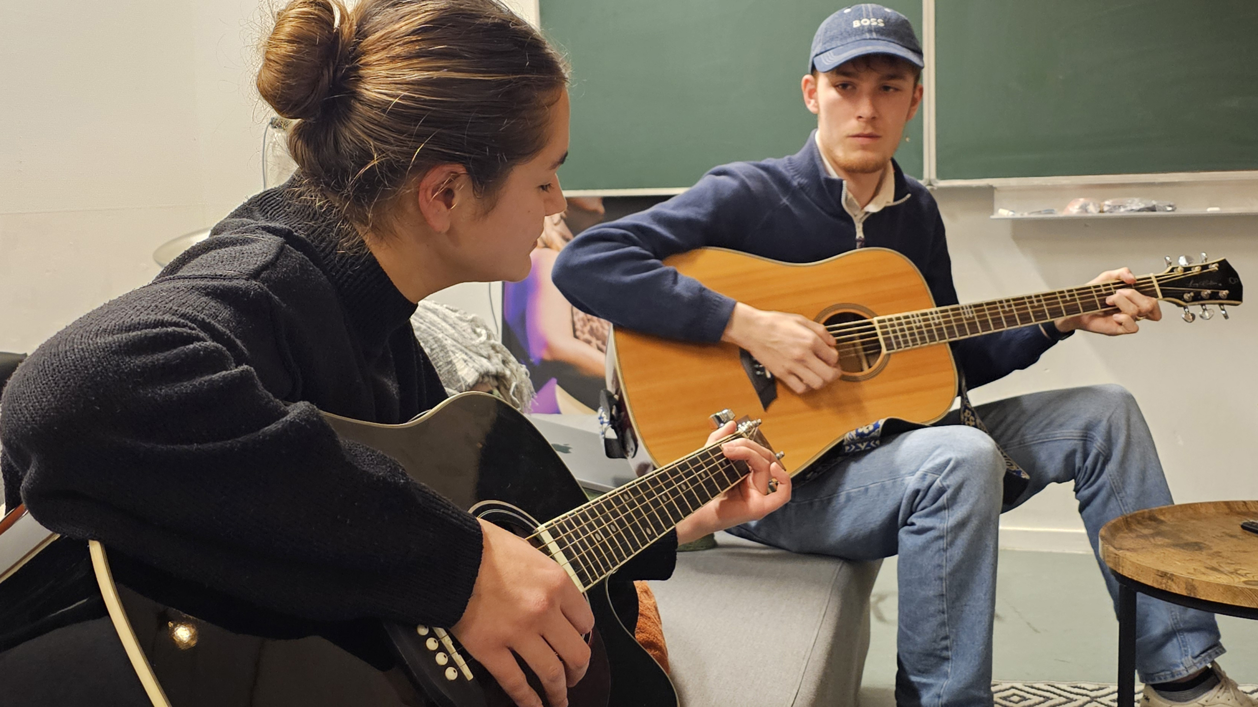 Twee mensen zitten binnen in een kamer en spelen elk op een akoestische gitaar, een vrouw met een zwarte gitaar en een man met een bruine gitaar; op de achtergrond is een schoolbord zichtbaar.