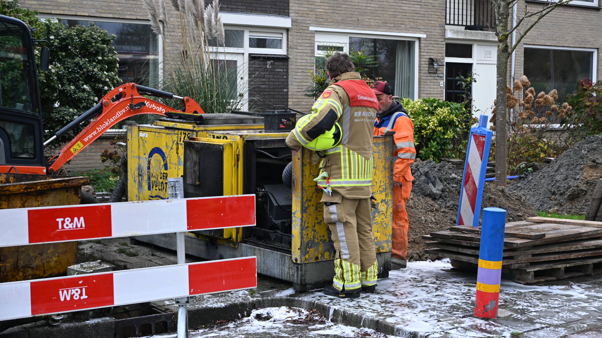 Twee mannen, waaronder een brandweerman in uitrusting en een arbeider in oranje veiligheidskleding, staan bij een gele generator op een bouwplaats voor een rijtjeshuis. Er is een rode graafmachine op de achtergrond en wegafzettingen met T&M-markeringen.
