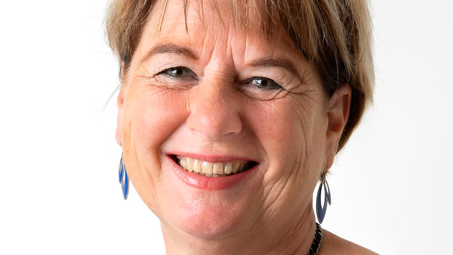 Vrouw met kort bruin haar en blauwe ogen, gekleed in een blauwe top, draagt een ketting met grote decoratieve bloemen.