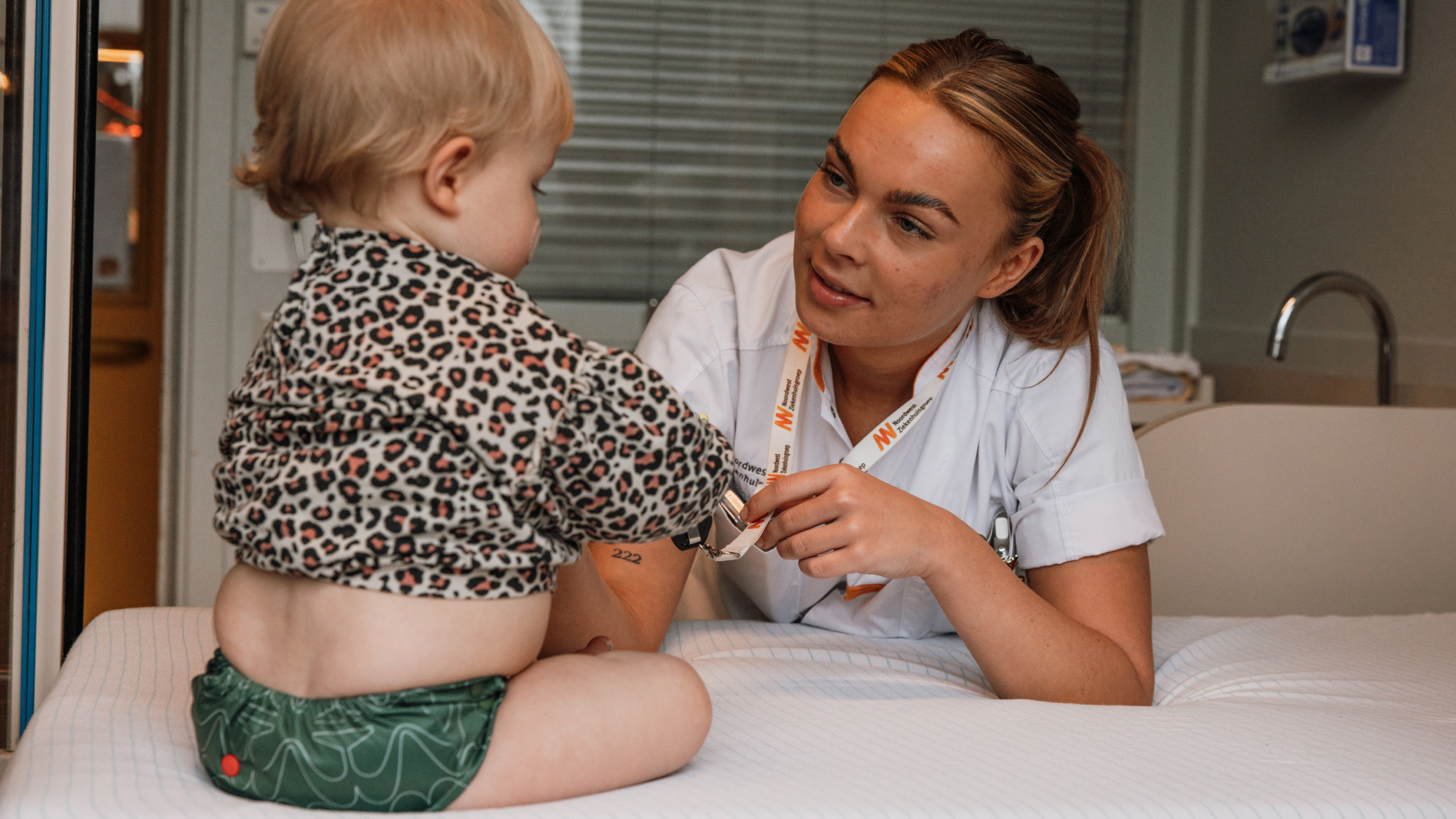 Een zorgverlener in witte uniformkleding onderhoudt een vriendelijke interactie met een jong kind in een ziekenhuisomgeving. Het kind draagt luierbroekjes en een trui met luipaardprint en zit op een bed.