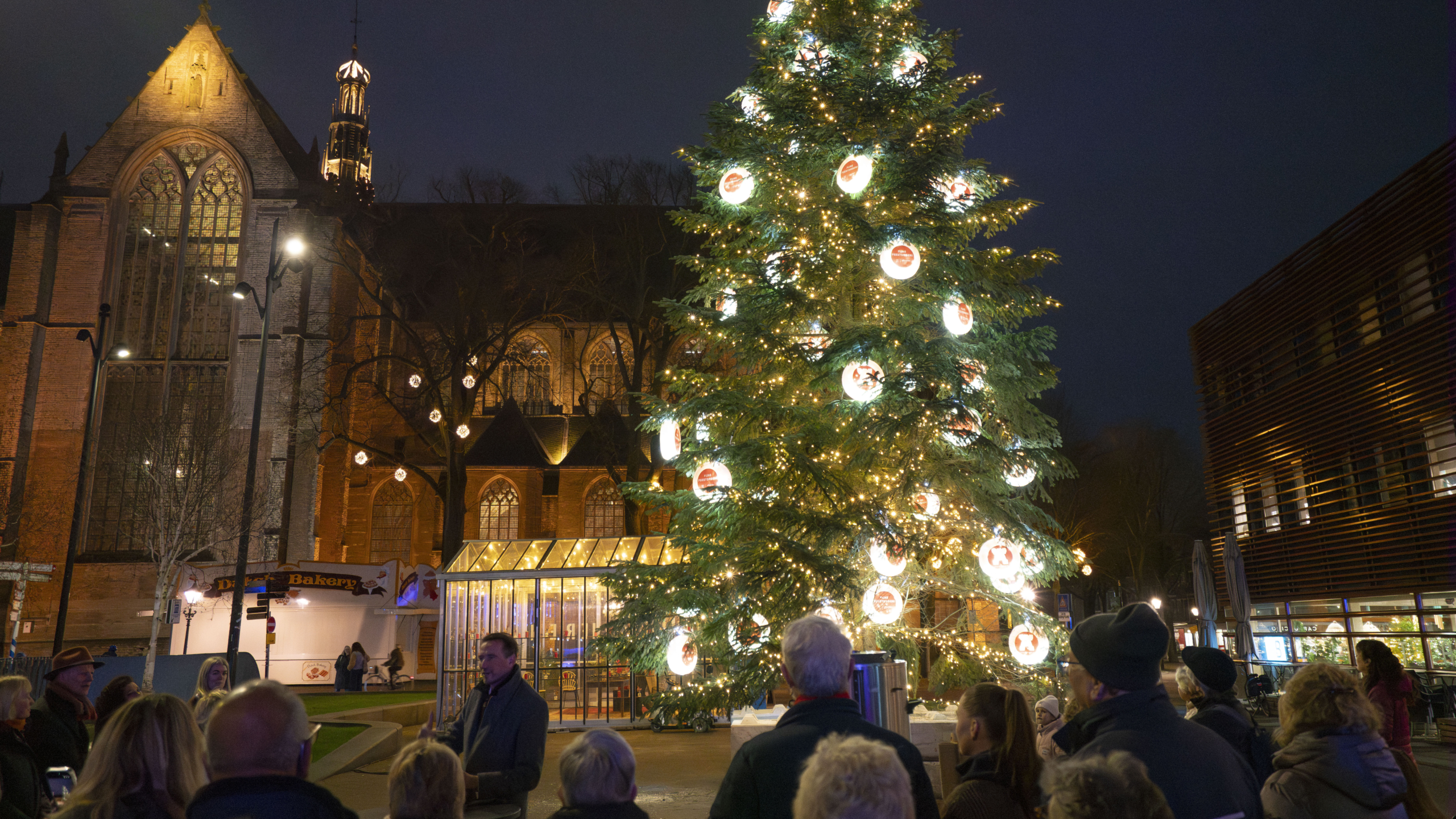 Verlichte kerstboom met versieringen voor een verlichte historische kerk, omringd door toeschouwers in de avond.