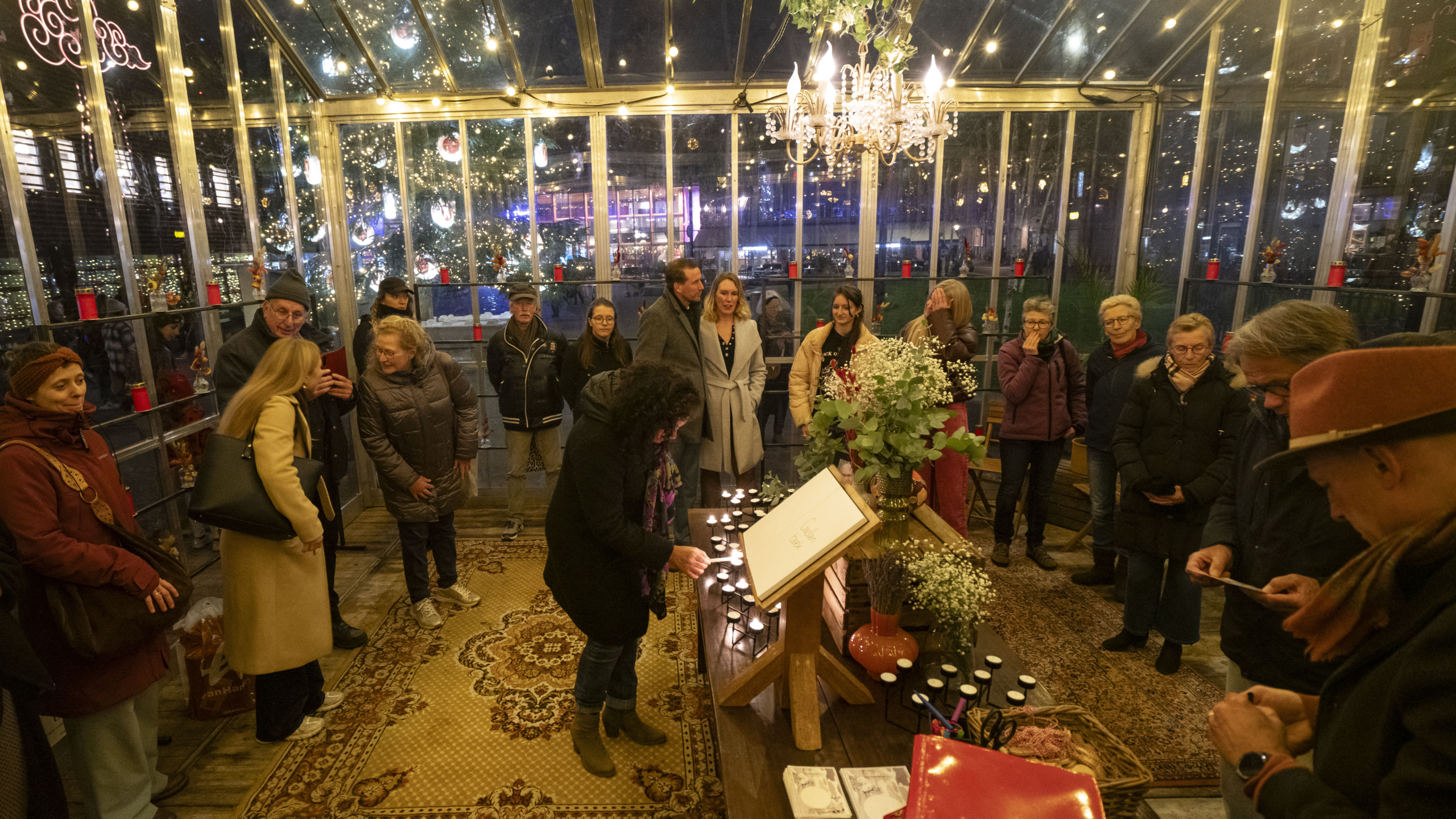 Een groep mensen verzameld in een verlichte serre tijdens een evenement, omgeven door decoraties zoals kaarsen en bloemen, met een kerstboom zichtbaar buiten in de achtergrond.