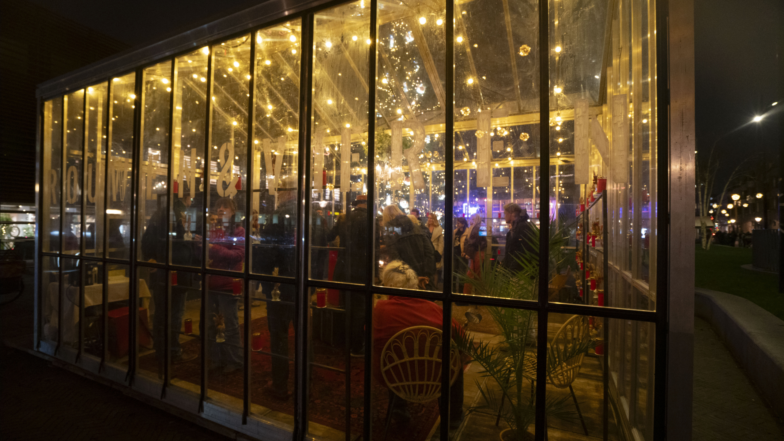 Mensen zitten en staan in een gezellig glazen paviljoen verlicht met warmgele lampjes, 's nachts in een stedelijke omgeving.
