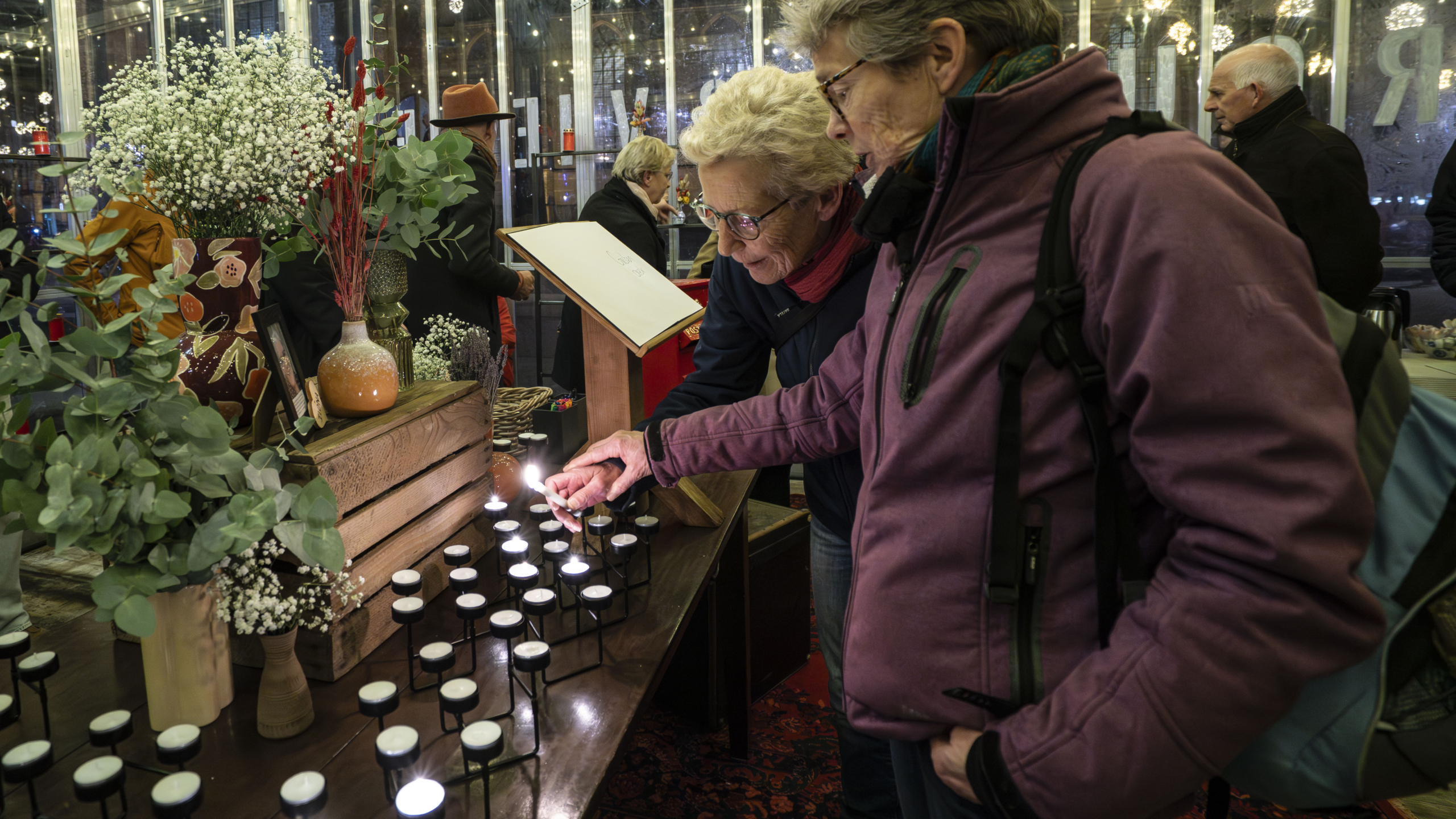 Twee oudere vrouwen met brillen steken kaarsen aan op een tafel met bloemen en decoratieve vazen, in een verlichte ruimte.