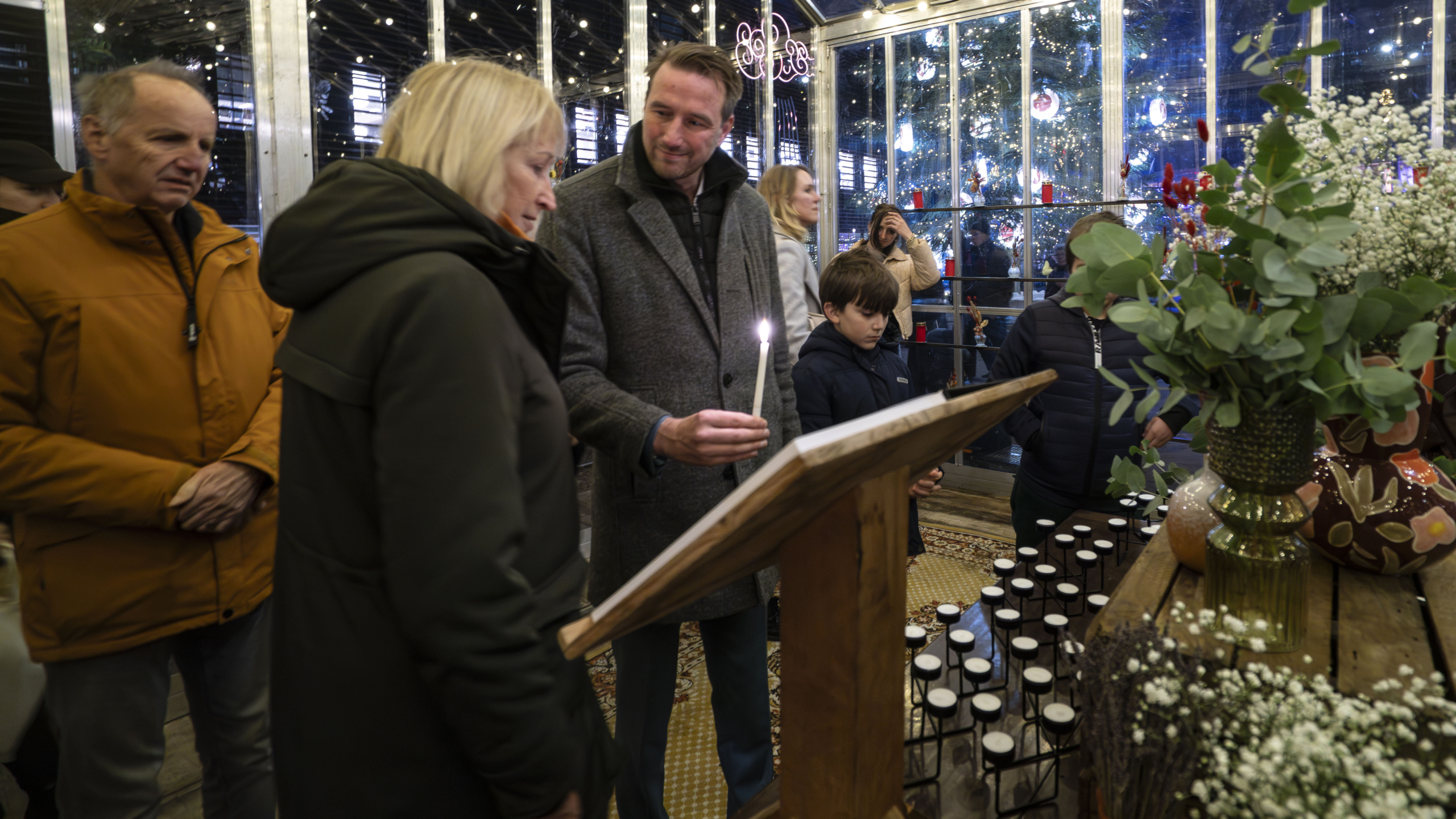 Mensen staan rondom een tafel met kaarsen en een bloemstuk, terwijl één man een brandende kaars vasthoudt in een ruimte met feestverlichting.