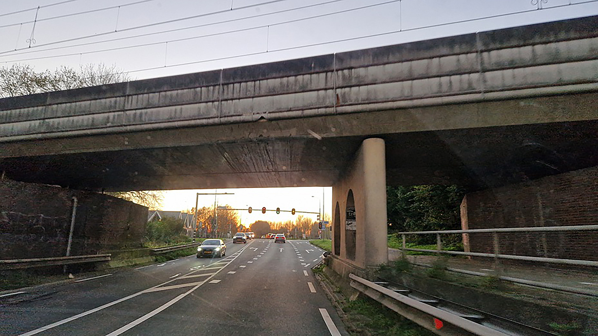 Verkeersweg met meerdere rijstroken die onder een betonnen viaduct doorloopt, met auto's op de weg en een stoplicht in de verte, tegen een achtergrond van avondlicht.