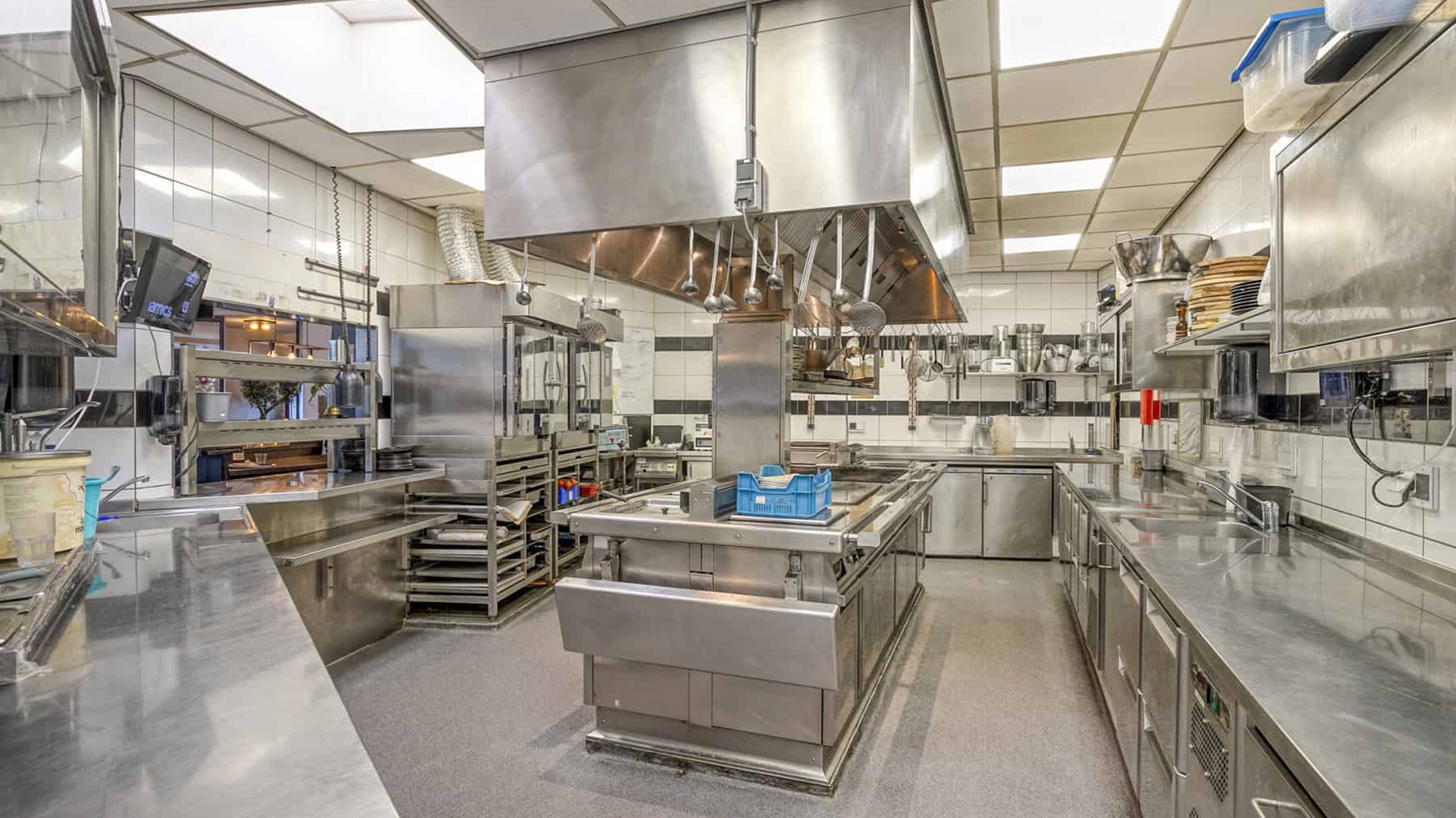 Professionele keuken met roestvrijstalen werkbladen, apparatuur en keukengerei, waaronder lepels en pannen, opgesteld in een nette en georganiseerde ruimte.