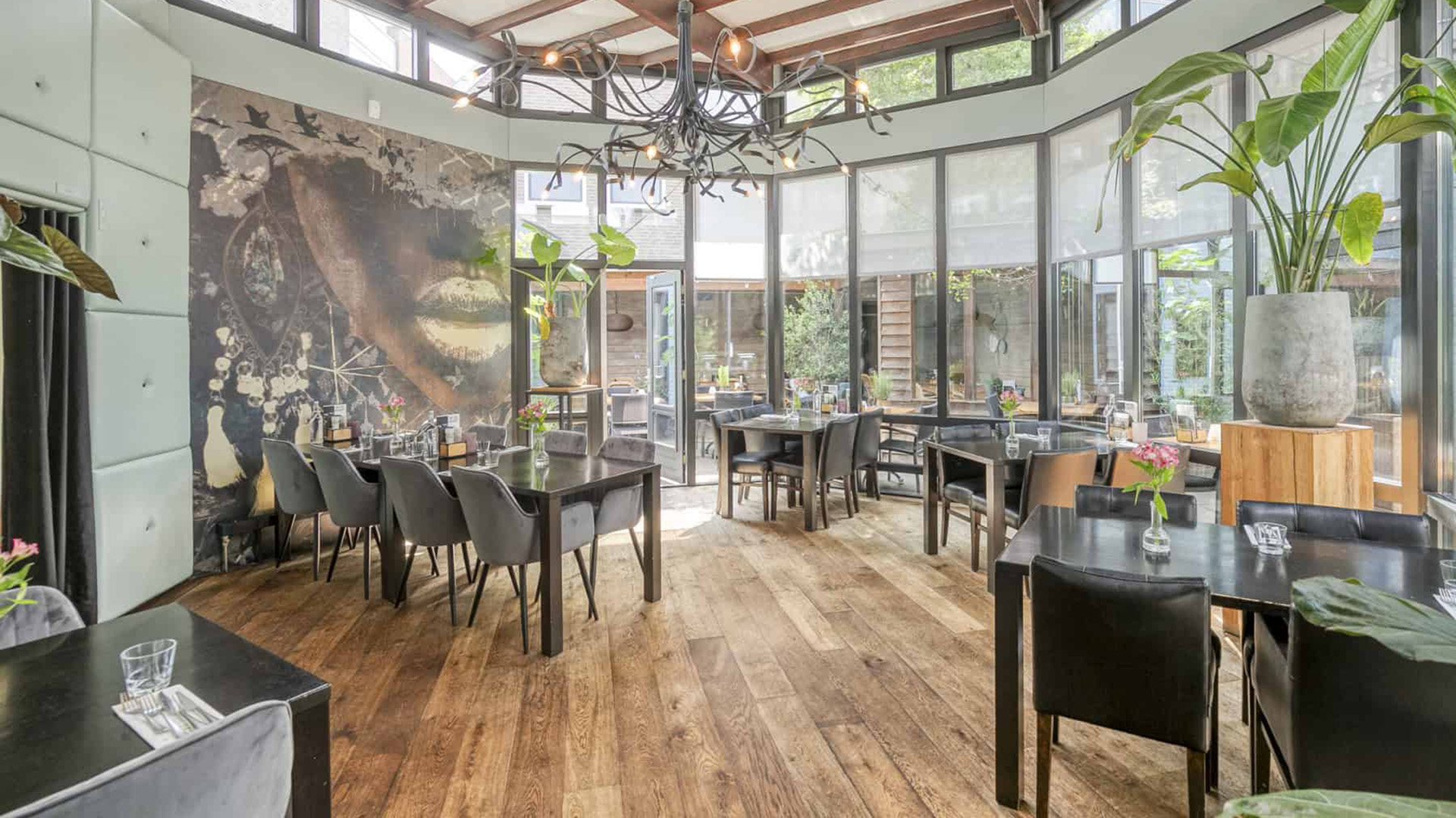 Een lichte, moderne eetruimte met houten vloeren, decoratieve planten, en glazen ramen, ingericht met zwarte tafels en stoelen omgeven door een grote muurschildering en een opvallende zwarte kroonluchter aan het plafond.