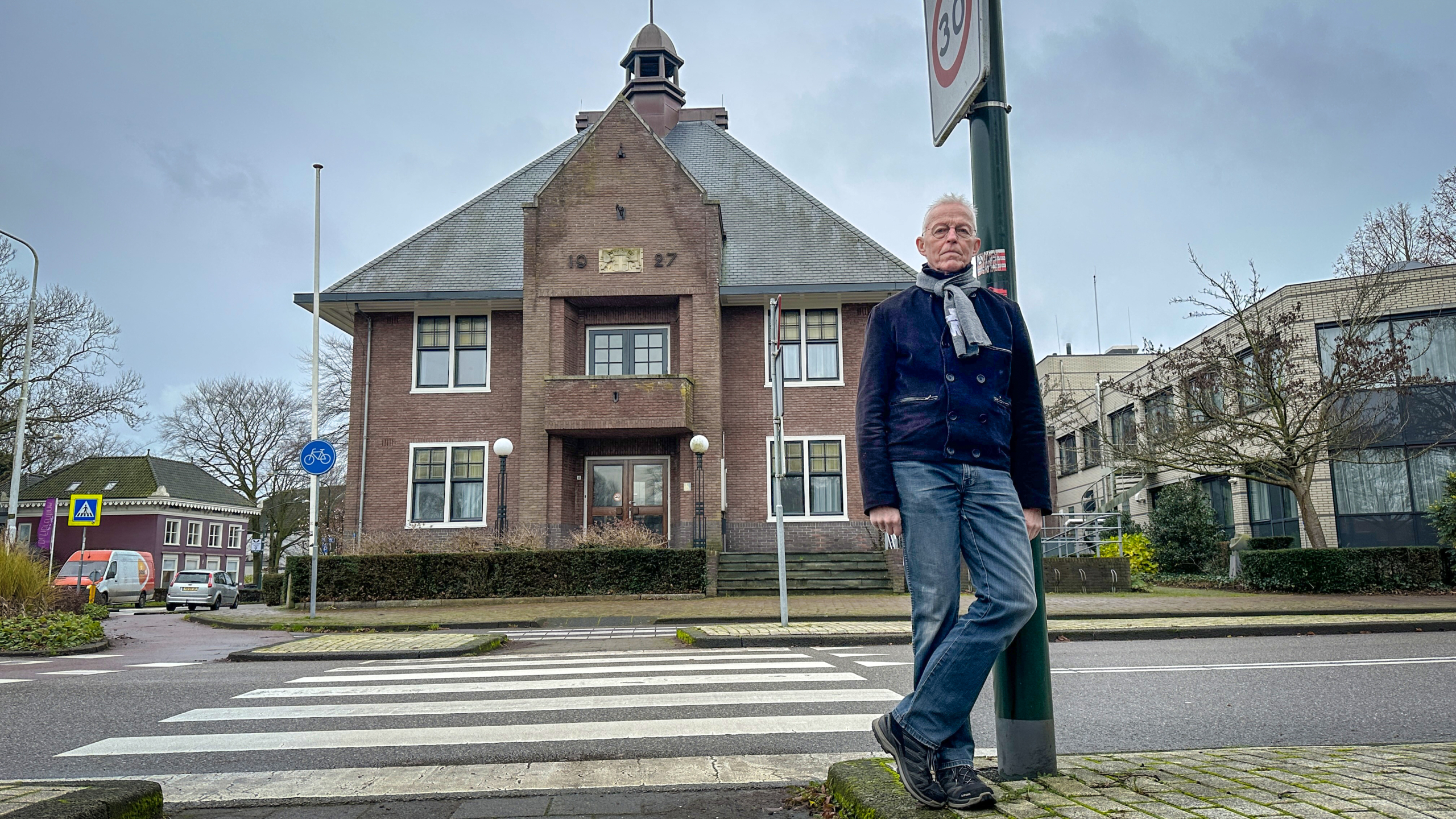 Een man leunt tegen een verkeersbord op een trottoir voor een historisch gebouw met het jaartal 1927 op de gevel.