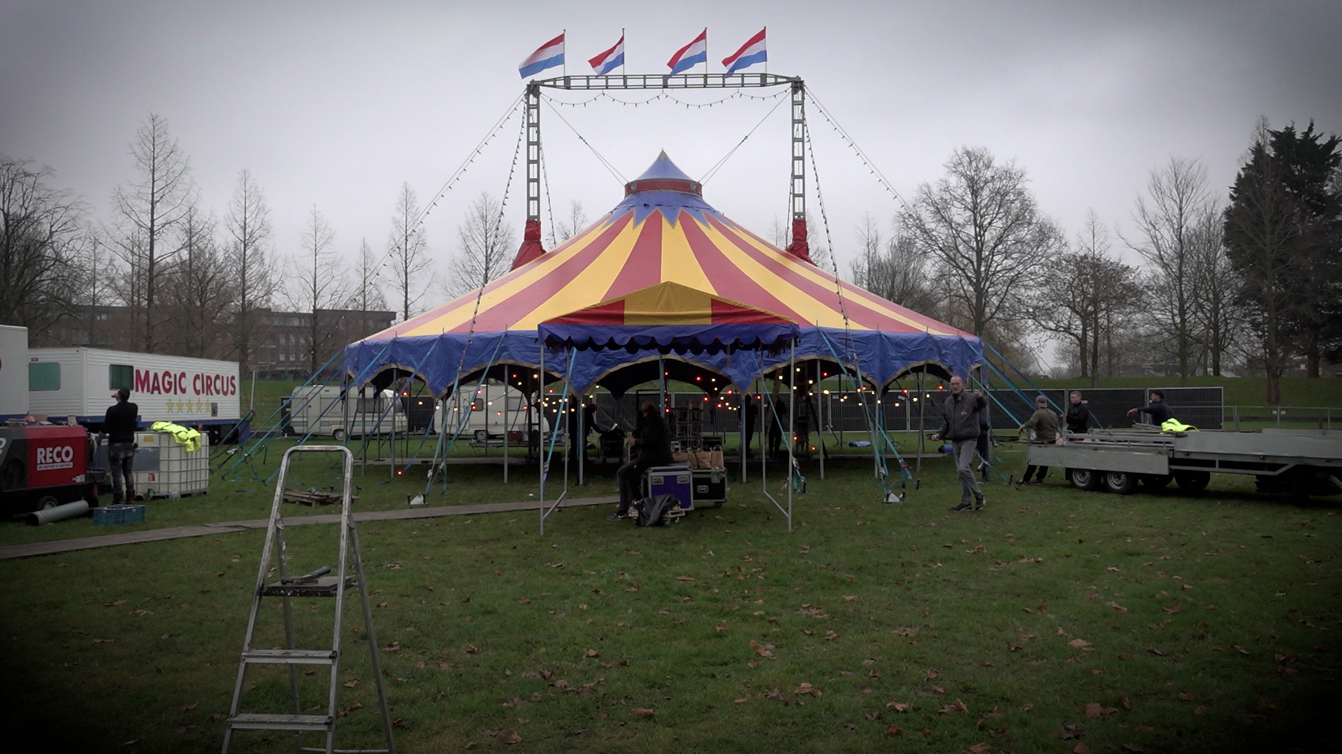 Een kleurrijke circustent wordt opgebouwd op een grasveld onder een bewolkte hemel, met Nederlandse vlaggen en enkele mensen die helpen bij de opbouw.