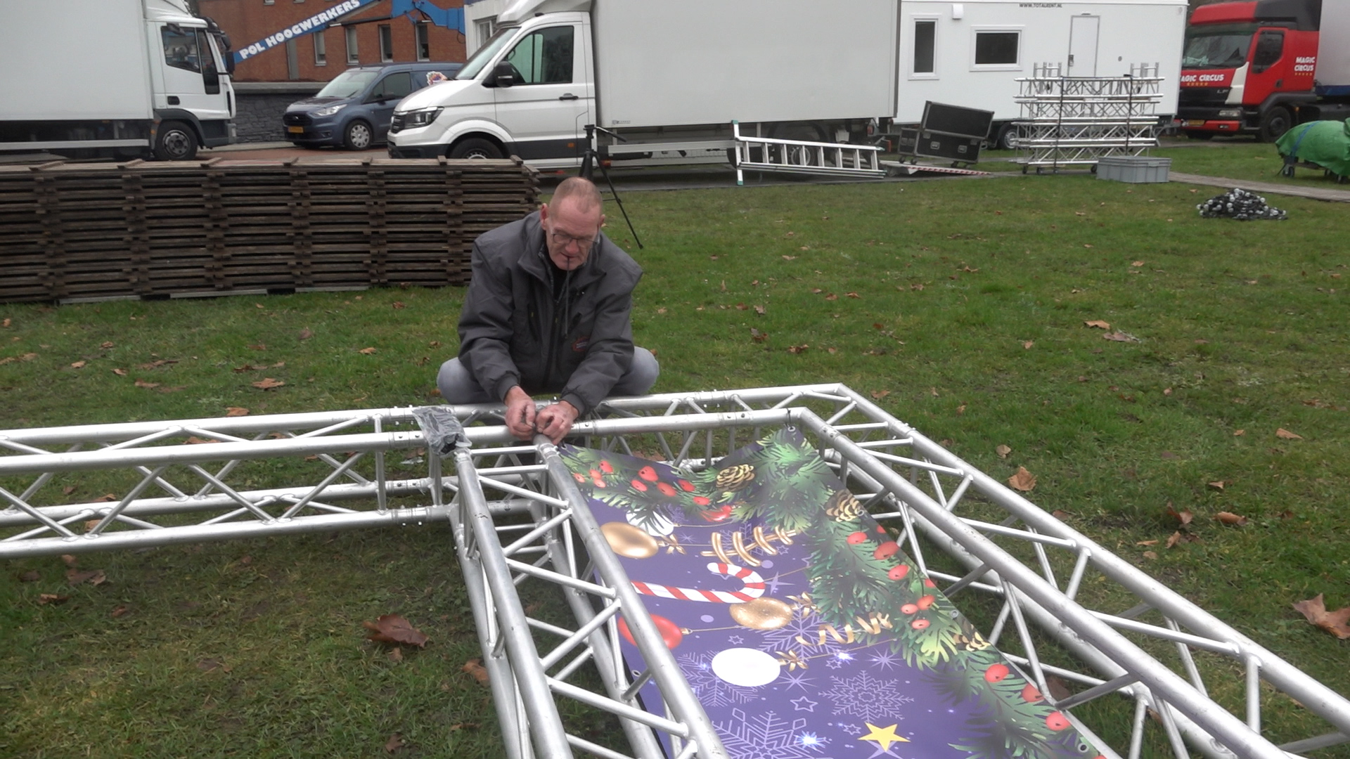 Man zet een decoratief spandoek vast aan een metalen truss-structuur op een grasveld, met geparkeerde voertuigen en gestapelde materialen op de achtergrond.