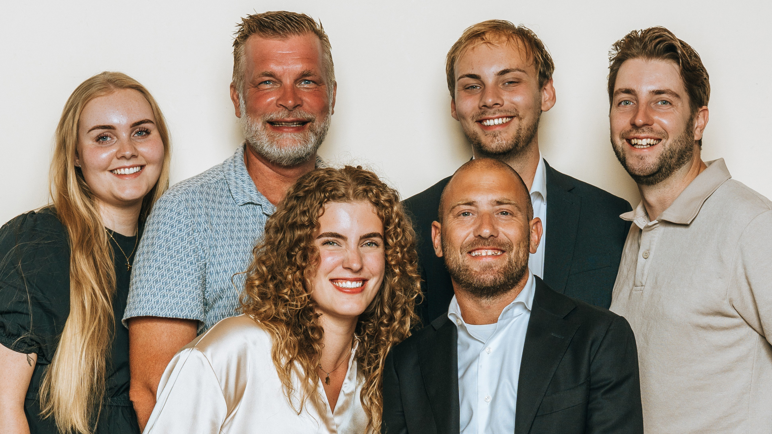 Groep van zes lachende mensen, bestaande uit vier mannen en twee vrouwen, poseren voor een neutrale achtergrond.