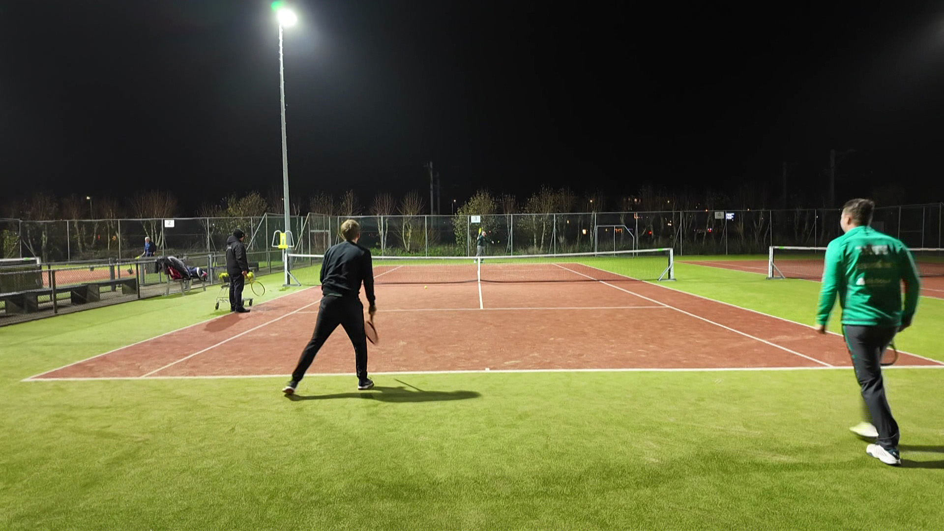 Mensen spelen tennis op een verlichte tennisbaan in de avond, geflankeerd door een omheining en enkele toeschouwers aan de zijkant.