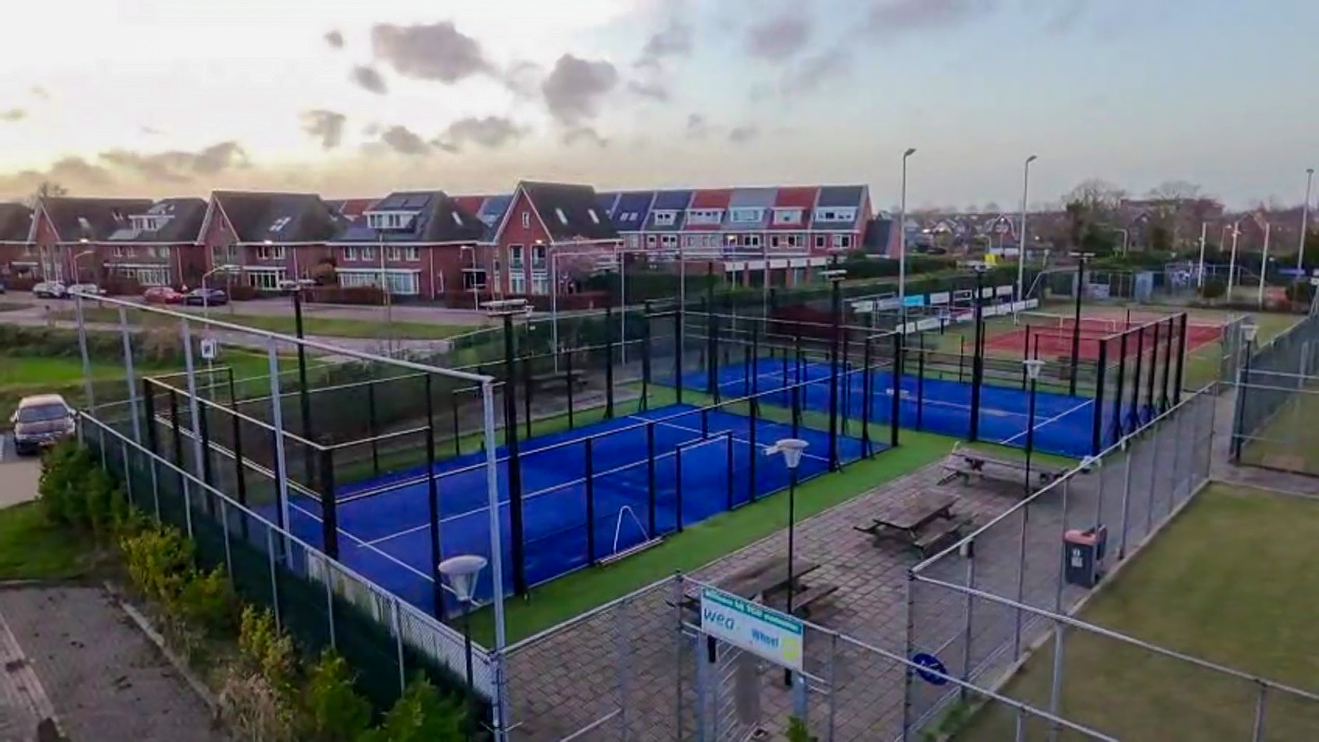 Padelbanen met blauwe ondergrond, omgeven door hekken, gelegen in een woonwijk met roodstenen huizen en auto's geparkeerd langs de straat.