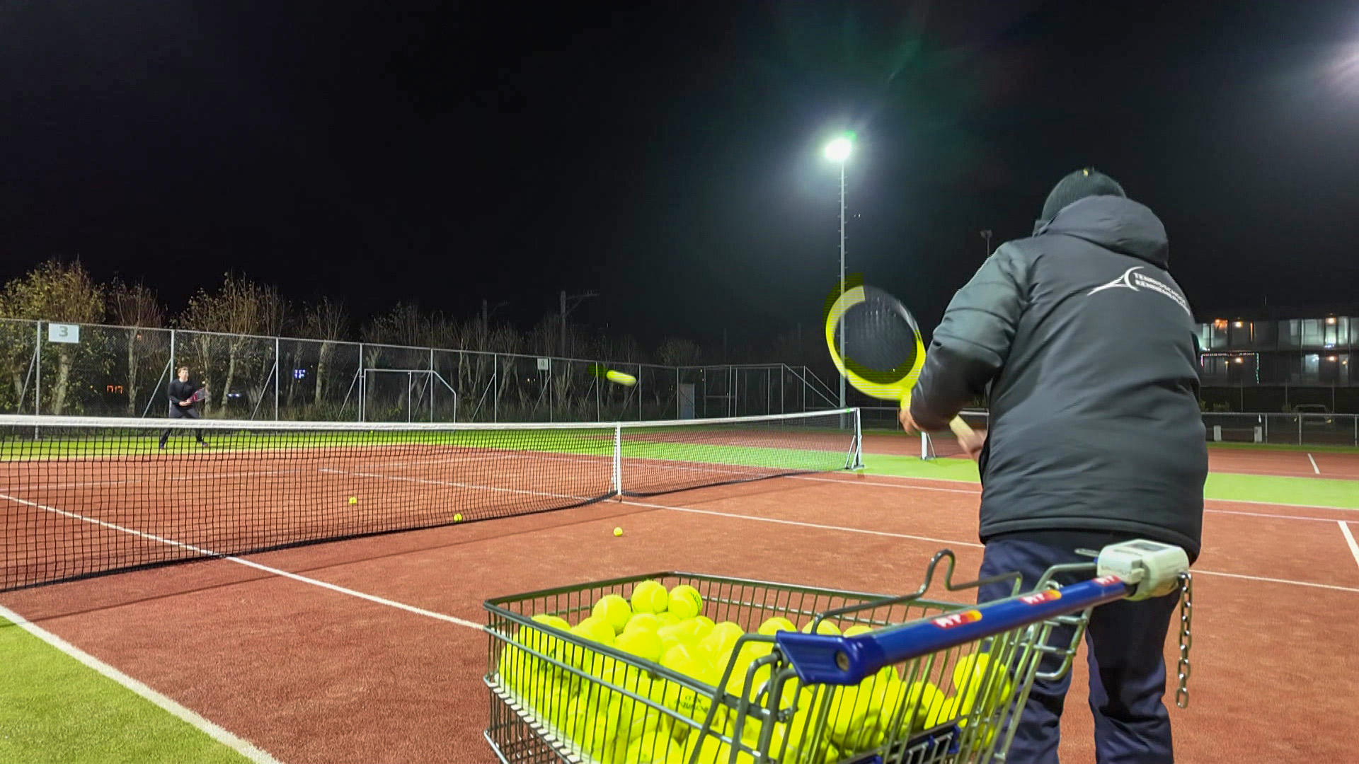 Twee mensen spelen tennis op een verlicht tennisveld in de nacht, terwijl een winkelwagentje vol met tennisballen op de voorgrond staat.