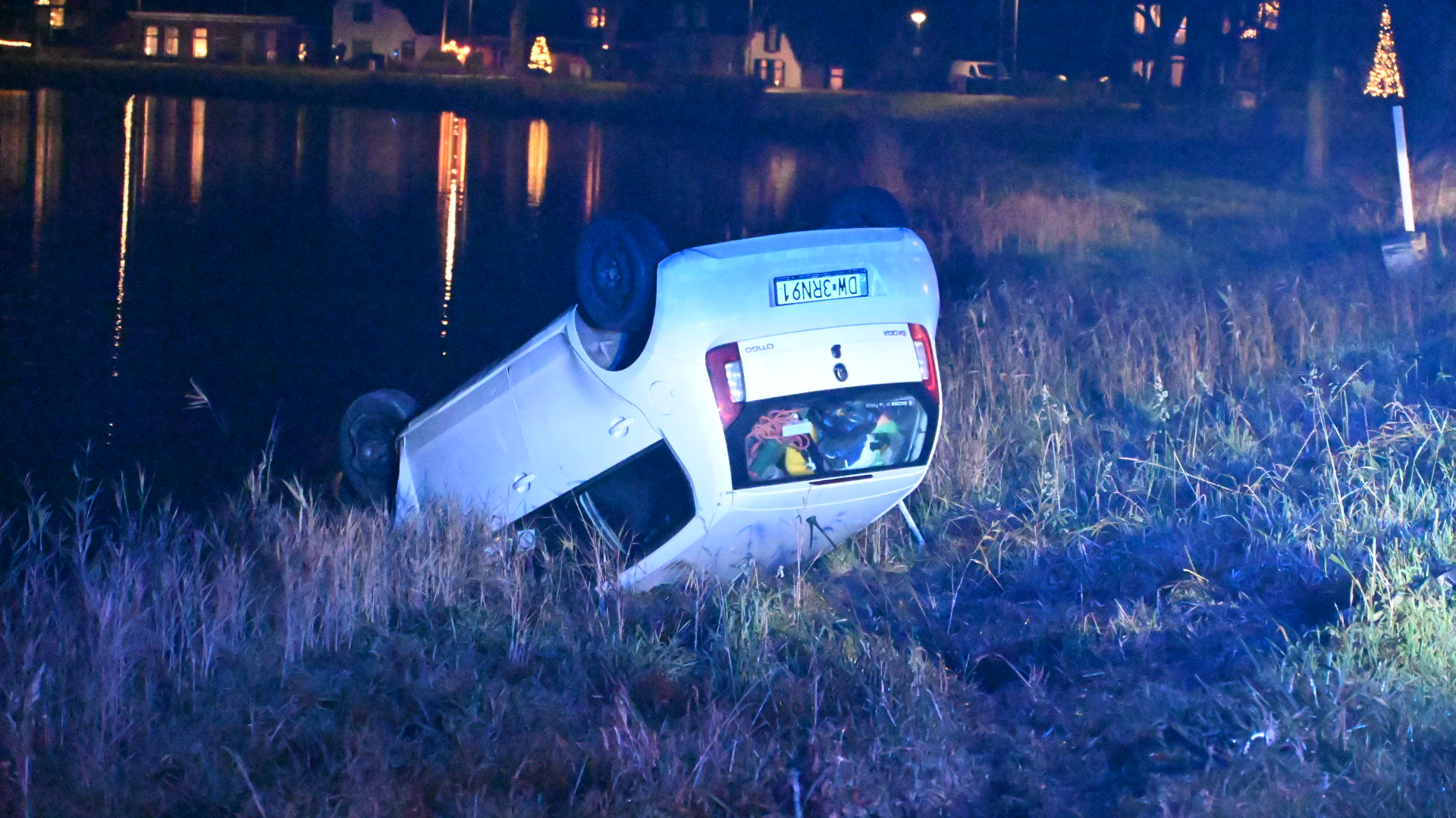 Auto op z'n kop in een sloot bij nacht, omgeven door gras en verlichte huizen in de achtergrond.