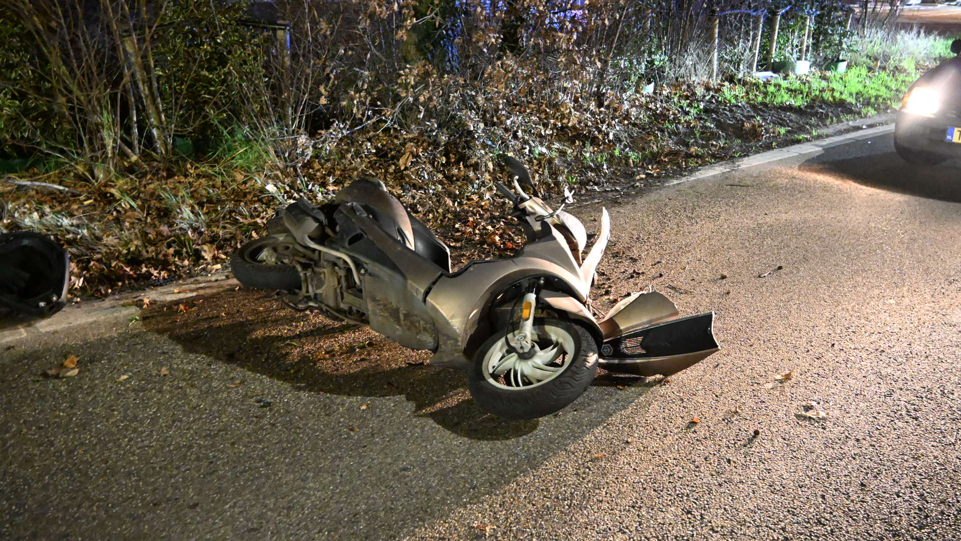 Een omgevallen scooter ligt beschadigd op een weg, omringd door bladeren en een auto aan de rechterkant.