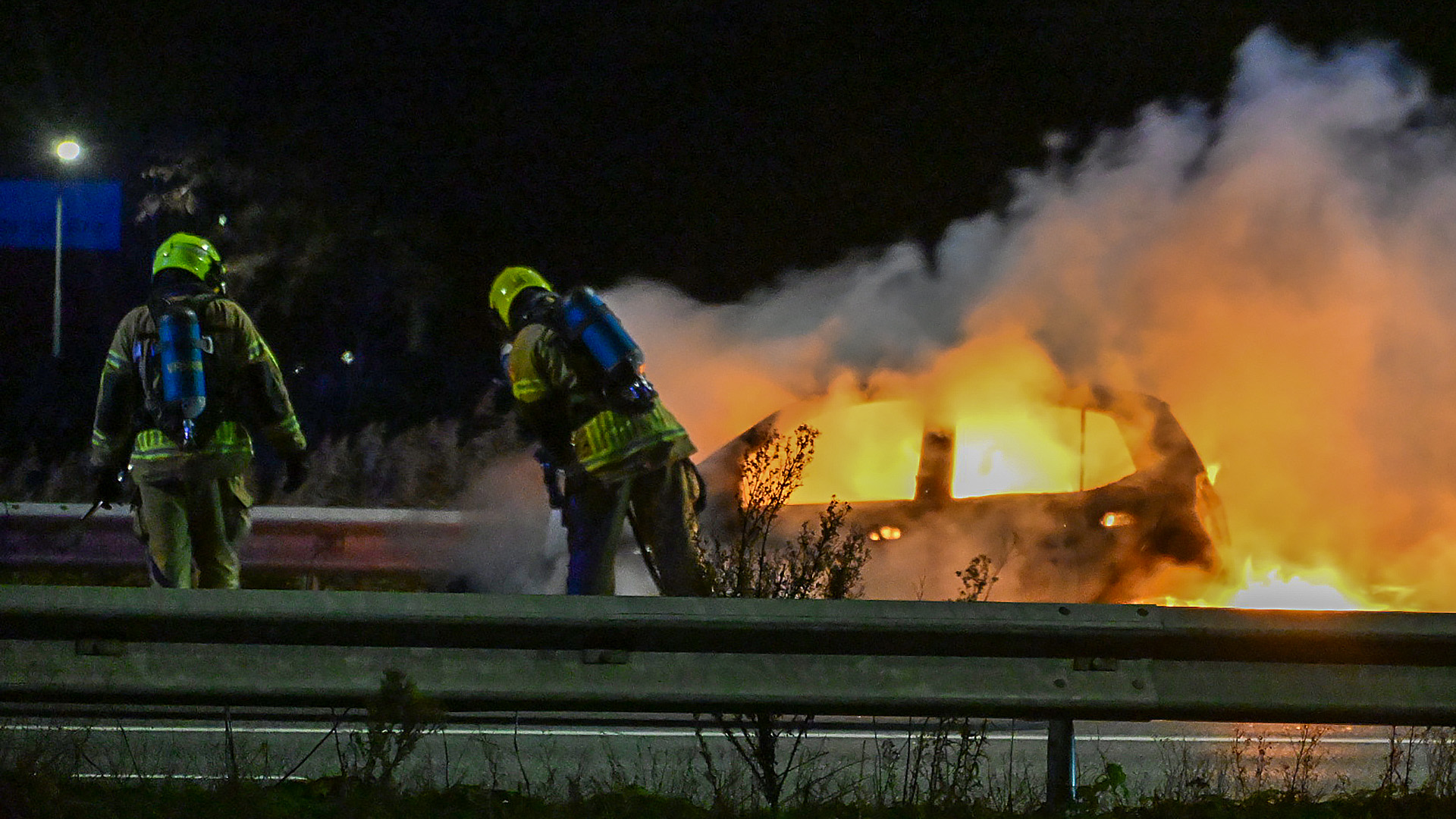 Twee brandweerlieden in beschermende kleding bestrijden een brandende auto langs de snelweg in het donker.