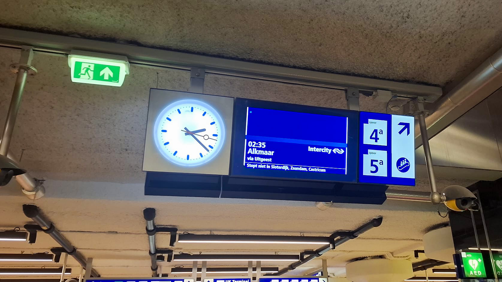 Stationinformatiebord met een klok die 2:27 toont, een vertrekbord voor een trein naar Alkmaar om 2:35 via Uitgeest, en aanwijzingen naar sporen 4a en 5a. Er is ook een nooduitgangsbord met een groene achtergrond en een wit pictogram van een rennend mannetje.