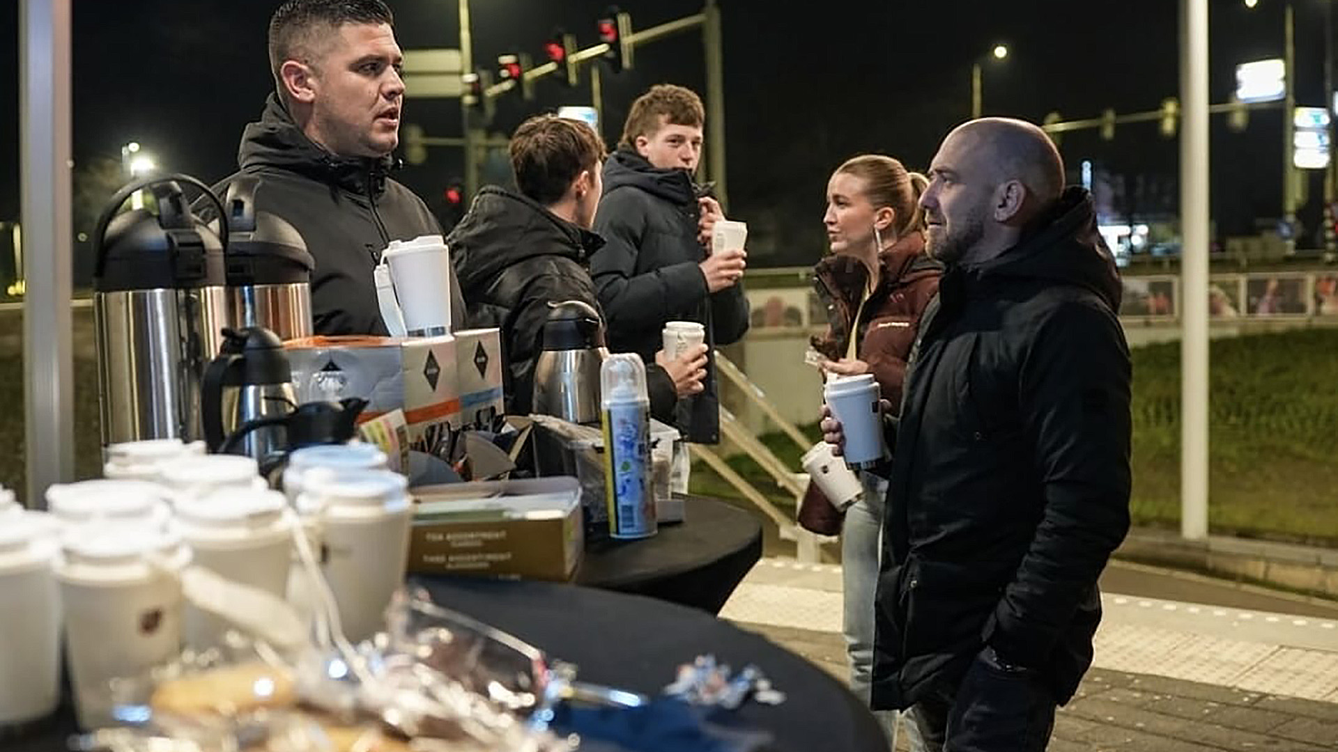 Een groep mensen in warme jassen staat 's nachts bij een tafel met koffie en snacks bij een kruispunt met verkeerslichten.