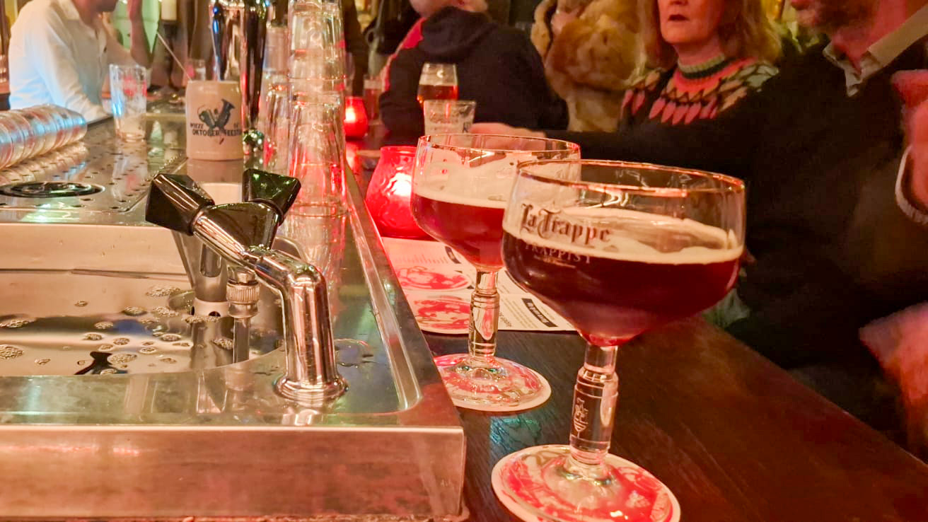 Een close-up van een bar met twee glazen bier, beide op een onderzetter met het logo van La Trappe Trappist, naast een tapkraan. Op de achtergrond zitten mensen aan de bar in zachte lichtverlichting.