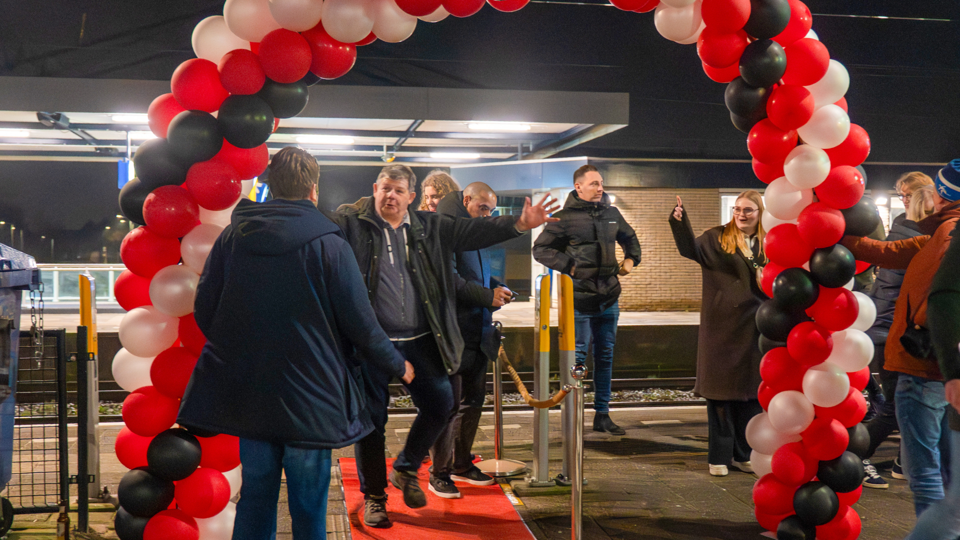 Mensen staan bij een boog van rode, witte en zwarte ballonnen op een perron, mogelijk tijdens een feestelijke gebeurtenis of opening.