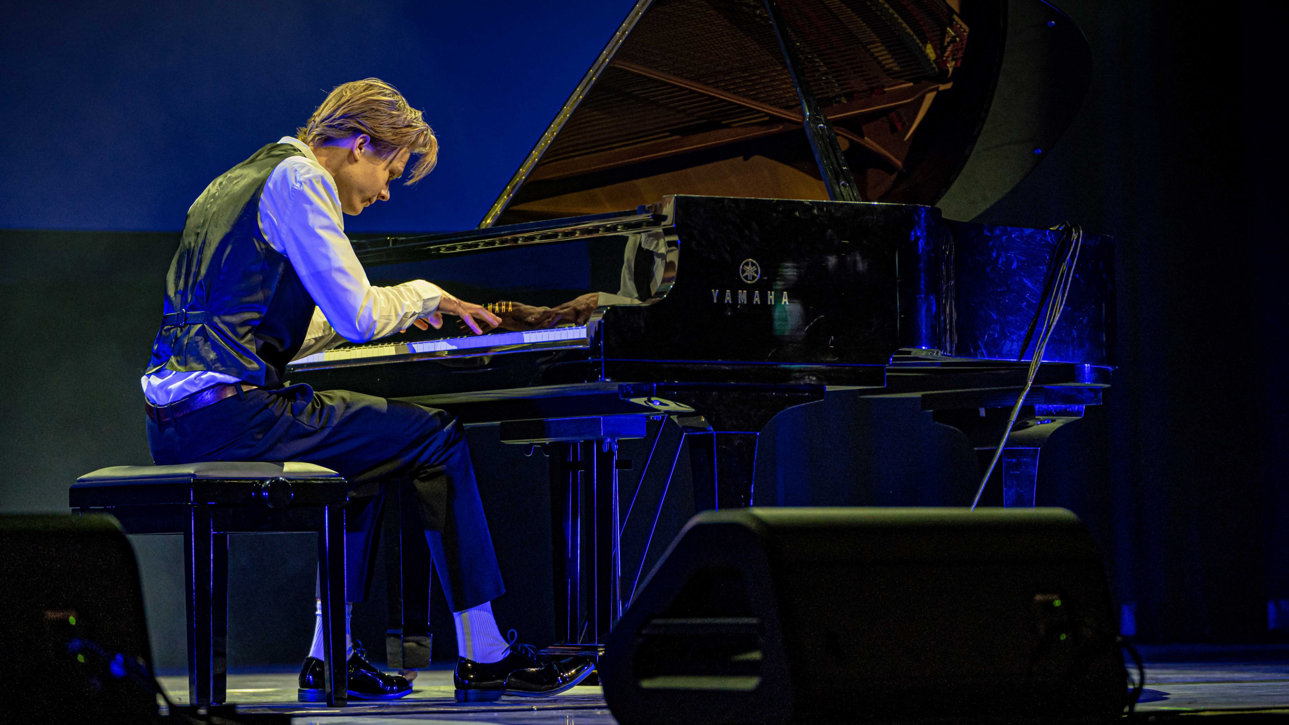 Een man in een vest en witte blouse speelt piano op een podium, onder blauwe belichting.
