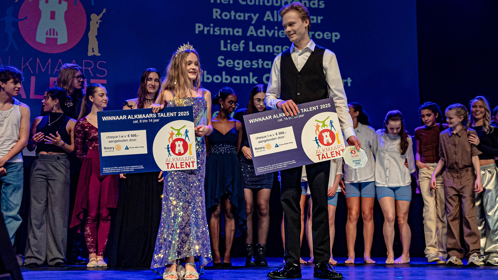 Twee jonge winnaars op het podium tijdens het Alkmaars Talent 2025 evenement, waarbij ze grote borden vasthouden met prijsinformatie. Op de achtergrond staan diverse deelnemers en supporters.