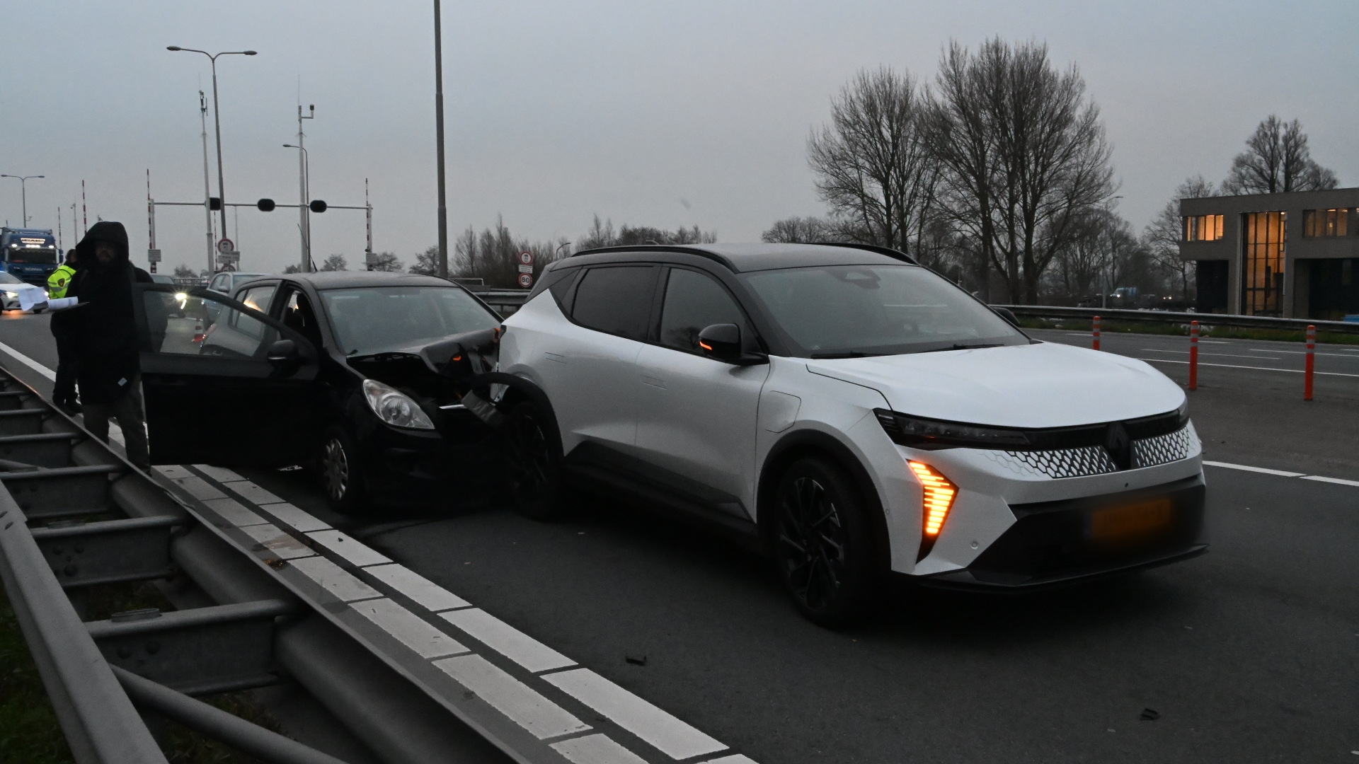 Een verkeersongeval op een weg, waarbij een zwarte auto en een witte SUV betrokken zijn; beide voertuigen zijn botsend tegen elkaar gekomen. De zwarte auto heeft zichtbare schade aan de voorkant. Er is een persoon in donkere kleding naast het incident, en een politieagent is in de achtergrond te zien.