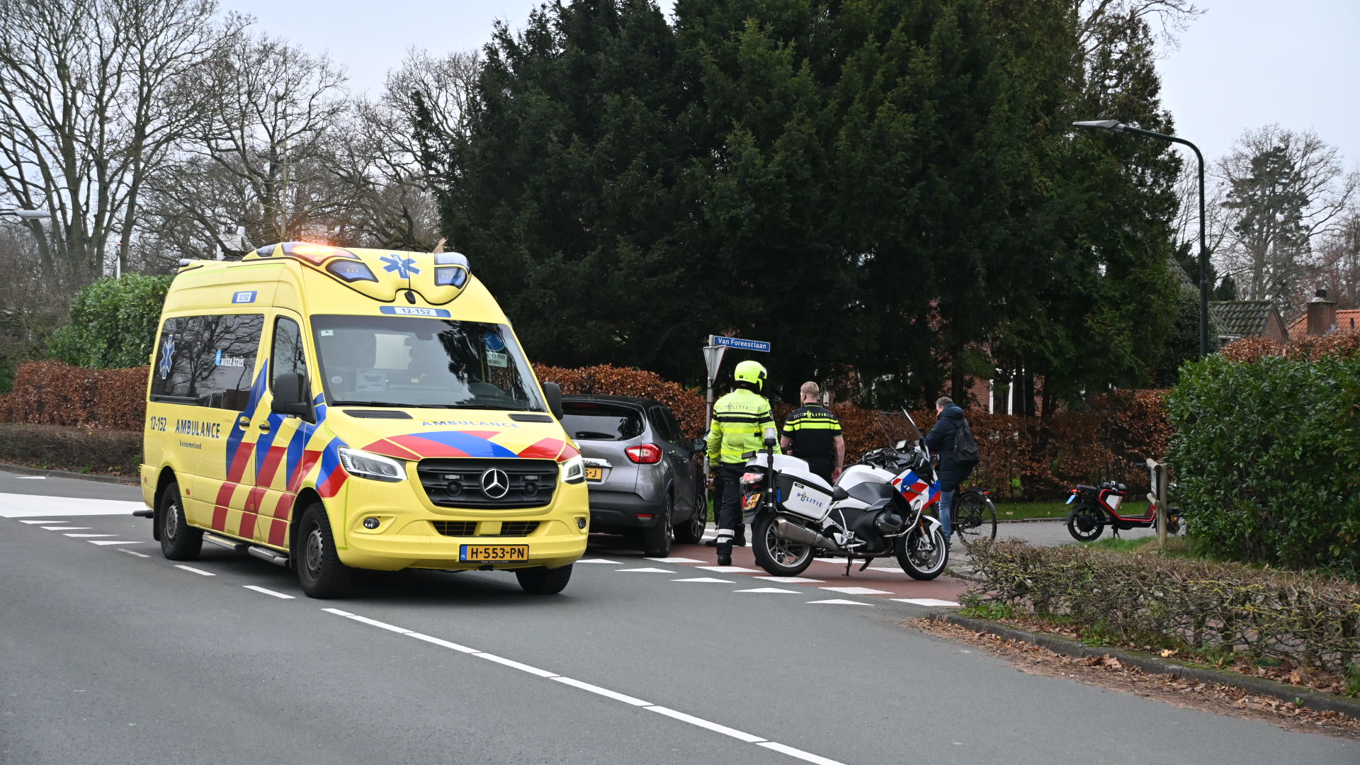 Een gele ambulance en een politieagent naast een motorfiets bij een kruising, met een auto en een fietser op de achtergrond, omringd door bomen en struiken.